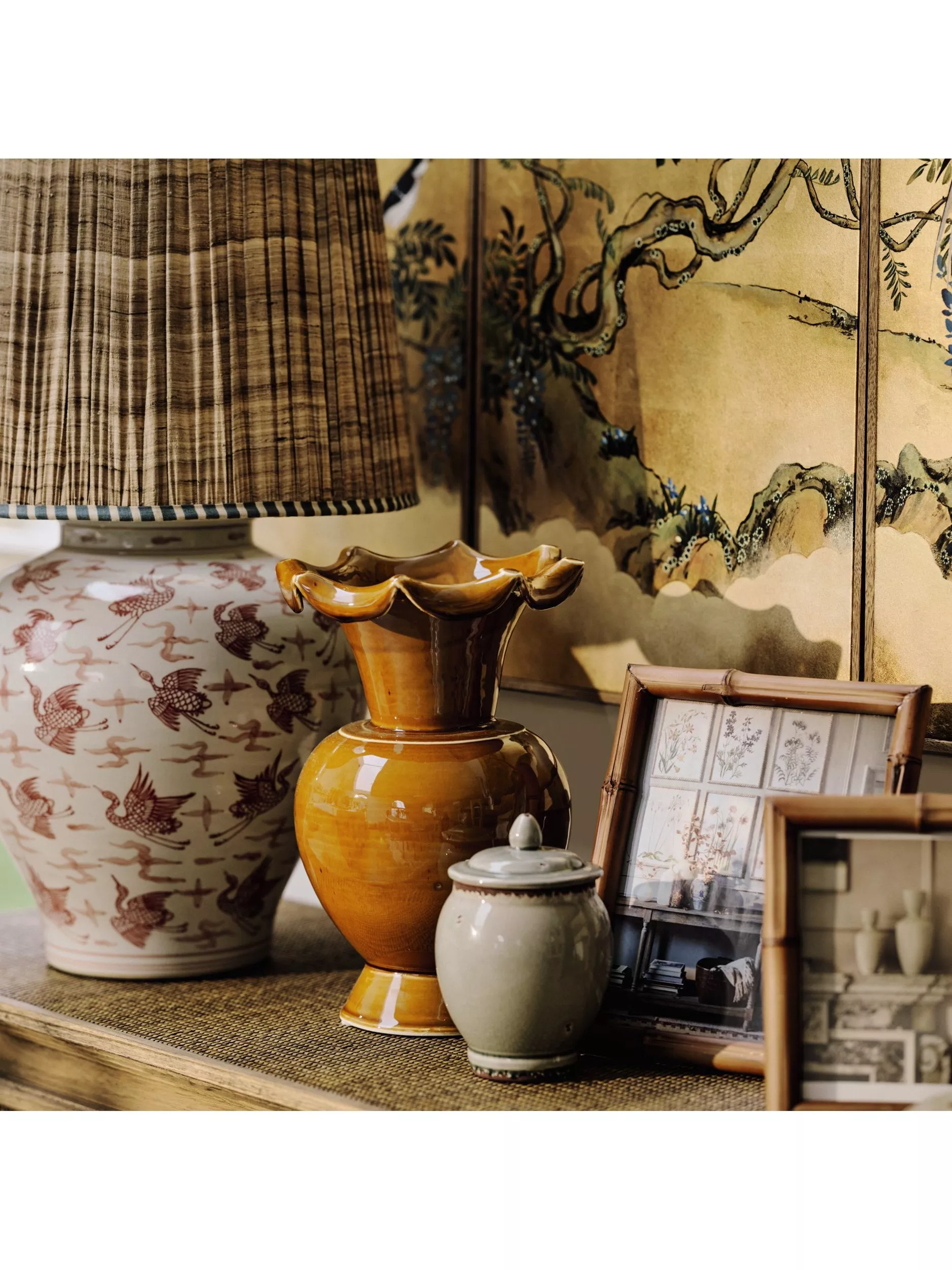 Vintage ceramic lamp and amber vase on sideboard with framed prints and oriental screen backdrop