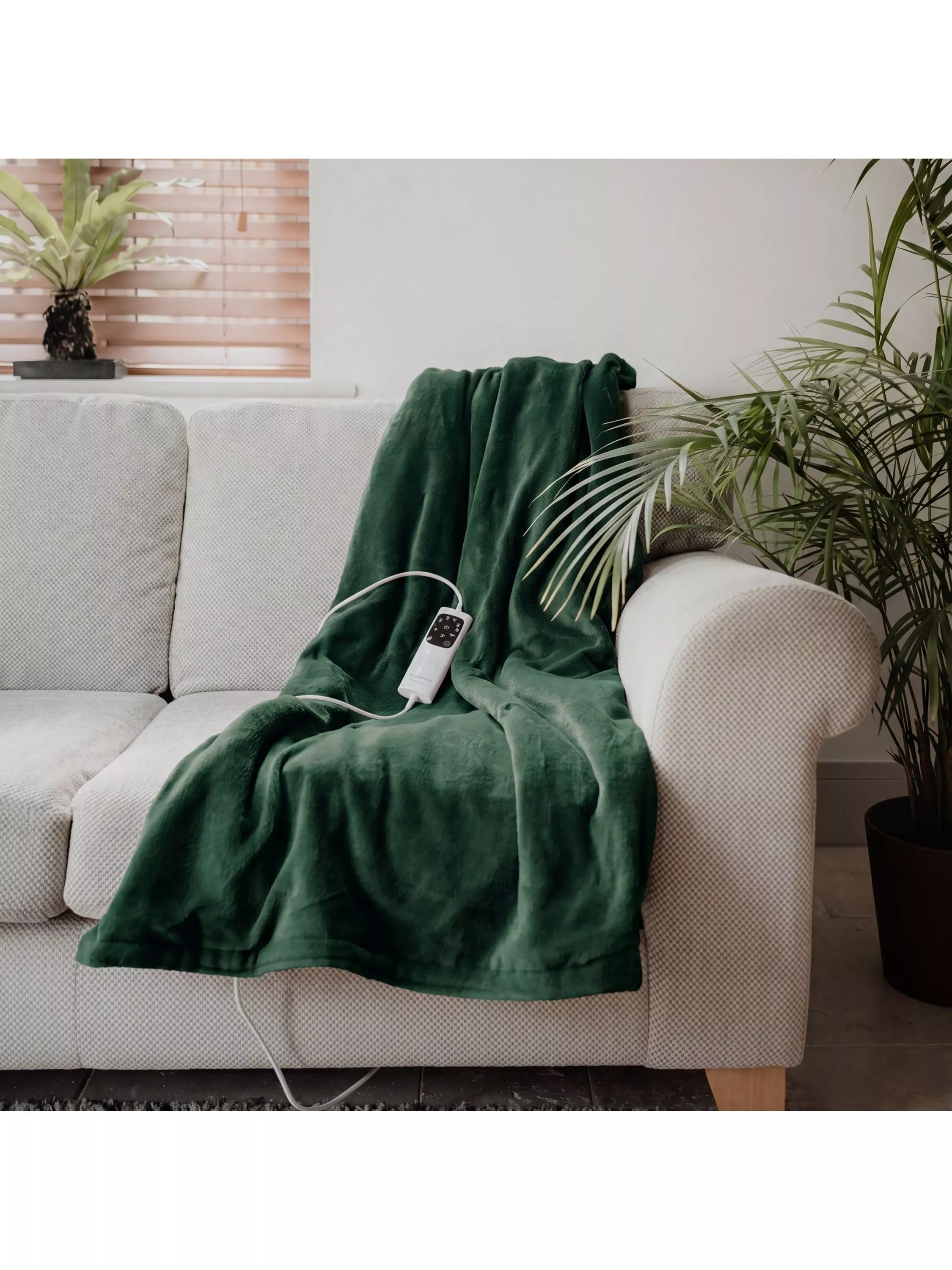 Green electric heated blanket with controller draped over beige sofa in modern living room with houseplants