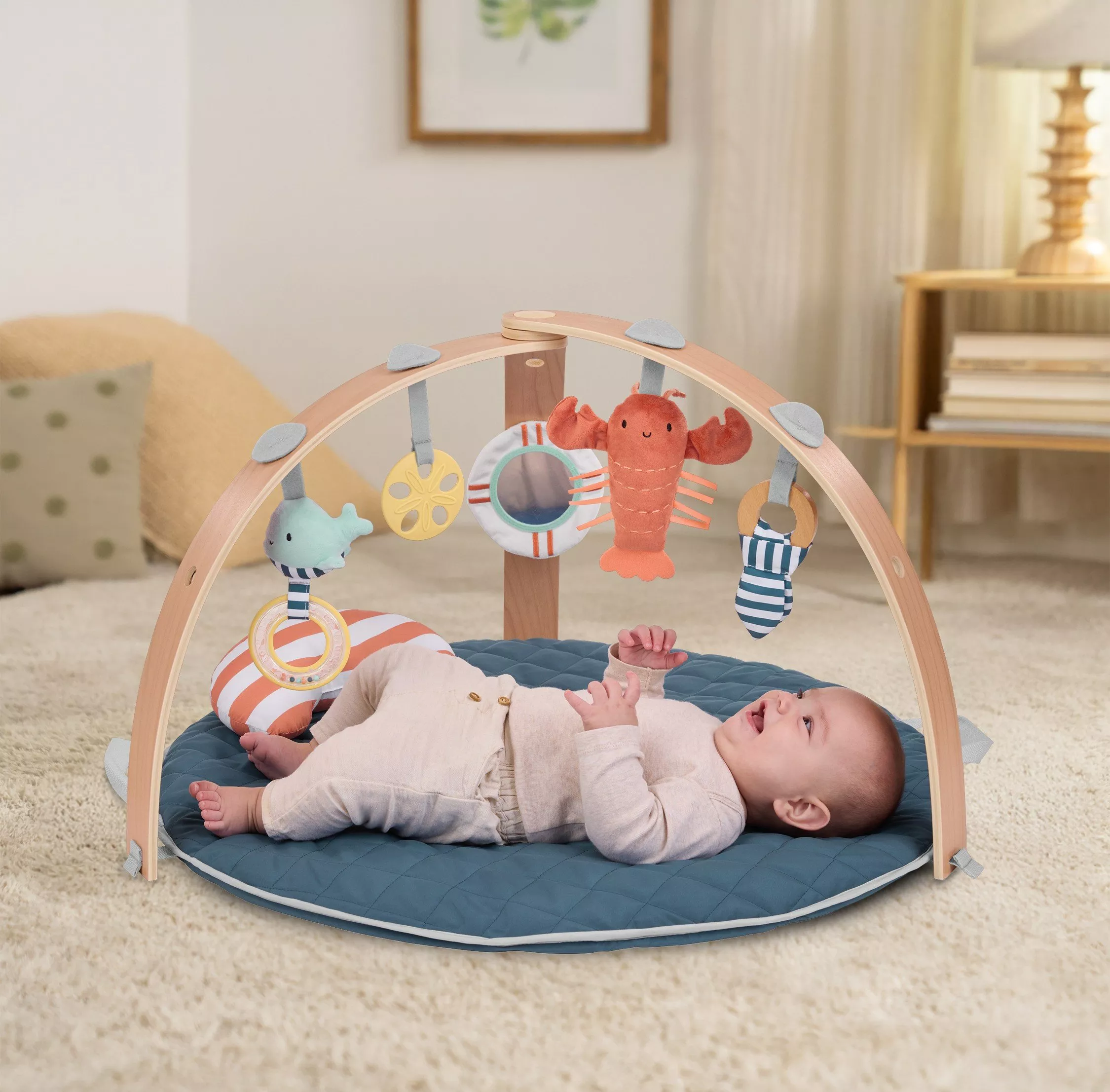 Baby lying on blue play mat under wooden activity gym with hanging sea creature toys in cosy living room