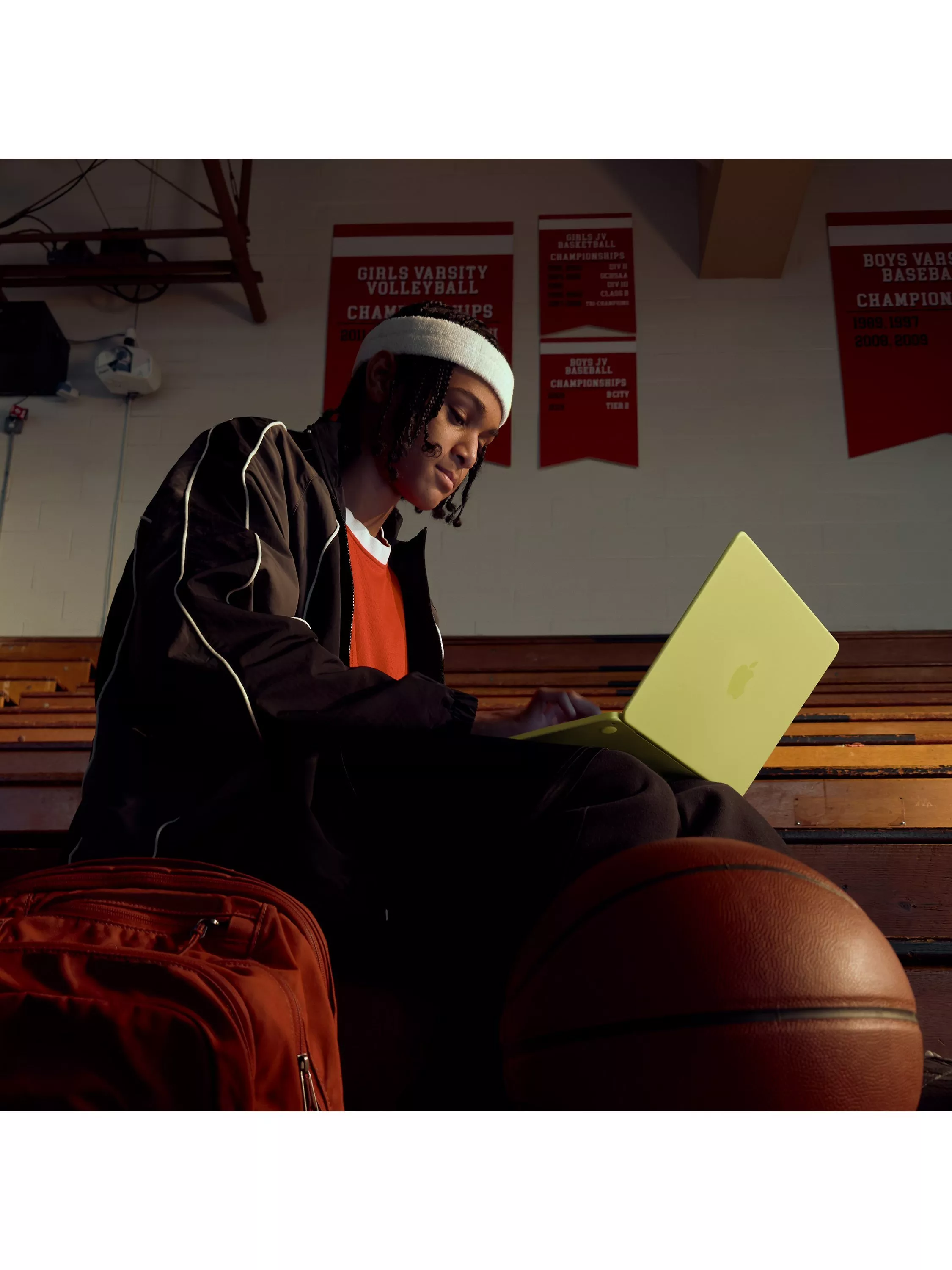 Student athlete using a laptop on gym bleachers beside basketball and backpack, with sports banners behind