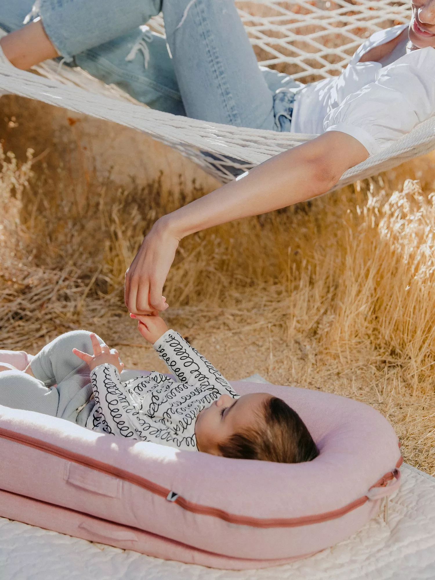 Mother relaxing in a hammock holding hands with baby lying on a pink lounger outdoors in dry grass