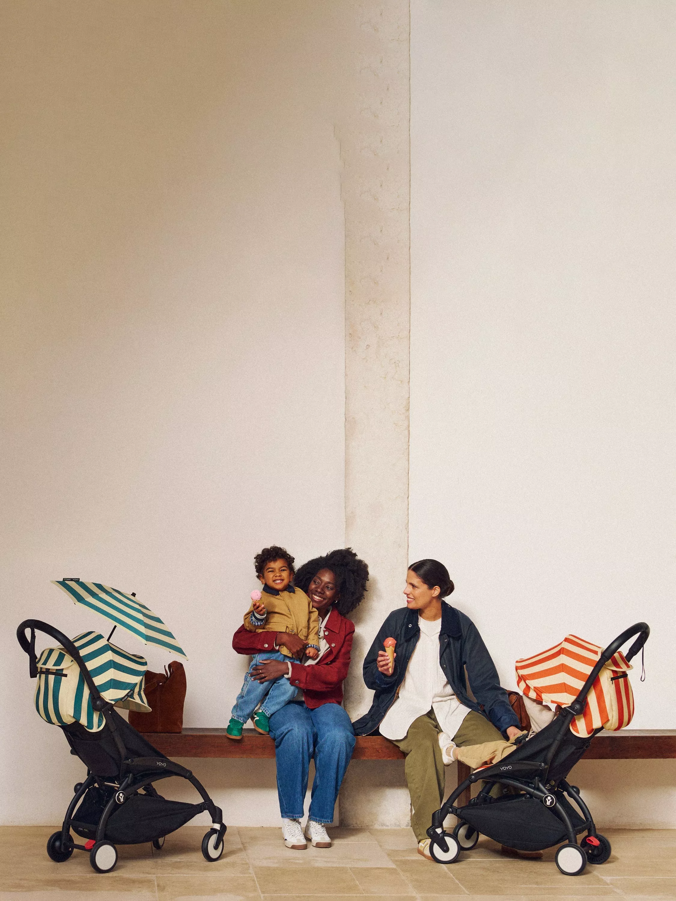 Two parents and toddler eating ice creams beside compact pushchairs with striped canopies, indoors on bench