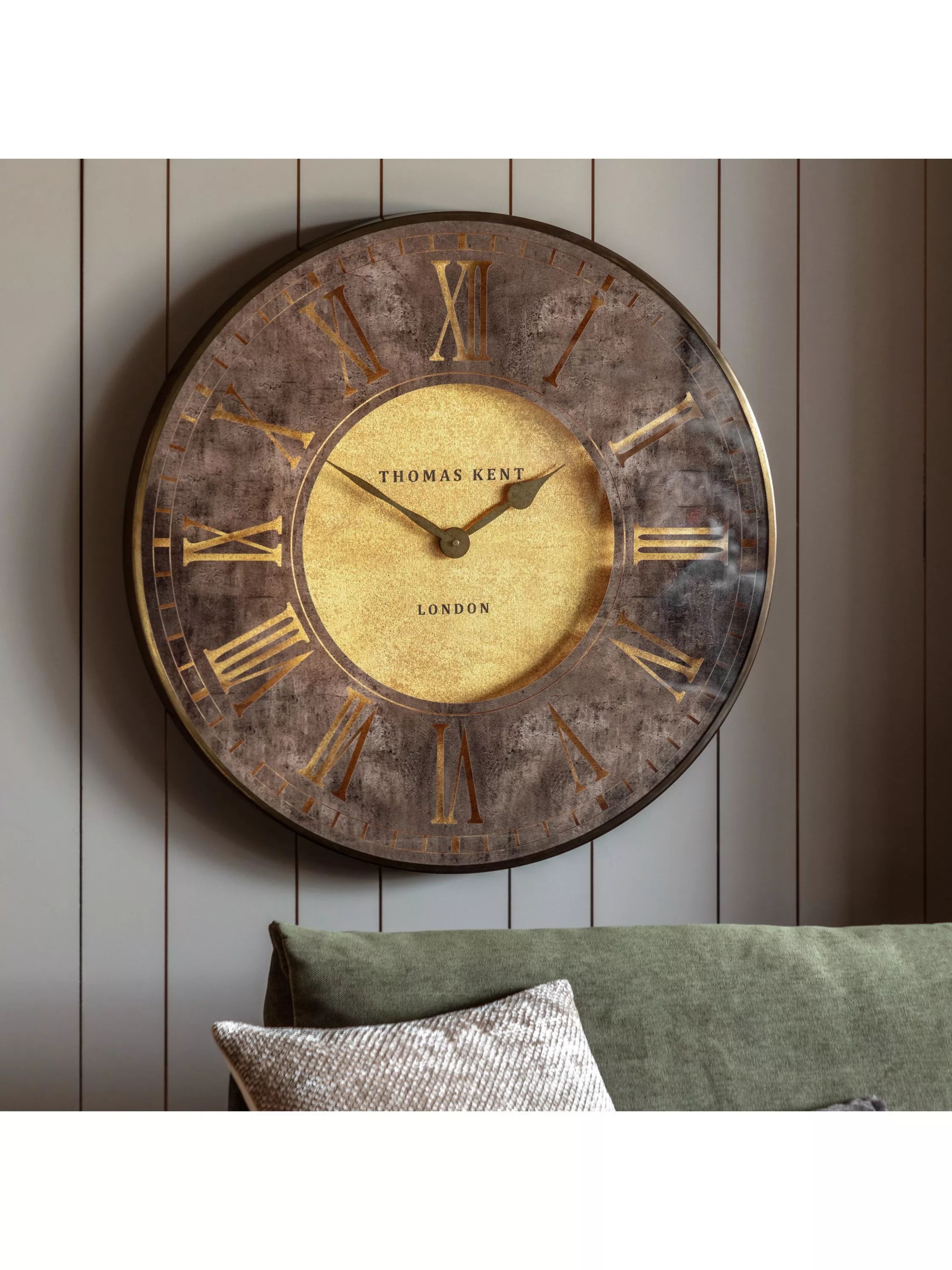 Large modern metal wall clock with Roman numerals above a wooden bench, cushion, throw and plant in hallway