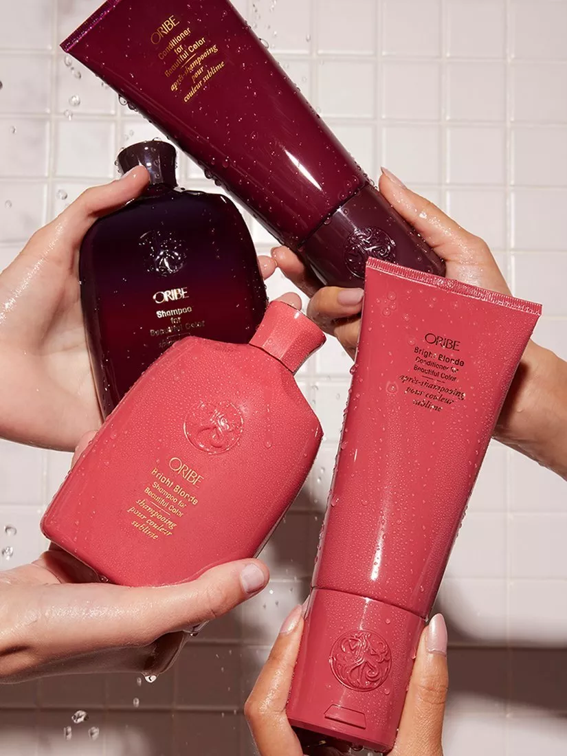 Oribe shampoo and conditioner bottles held in wet hands, colour care hair products against tiled shower wall