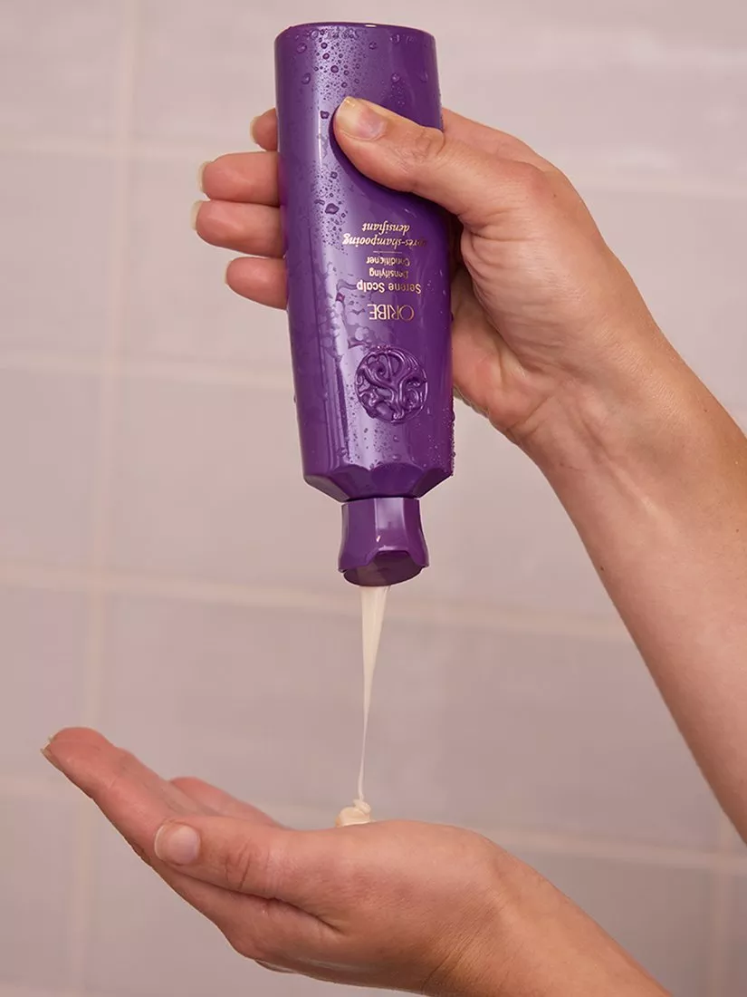 Hands dispensing Oribe Serene Scalp anti-dandruff shampoo from purple bottle in shower bathroom setting