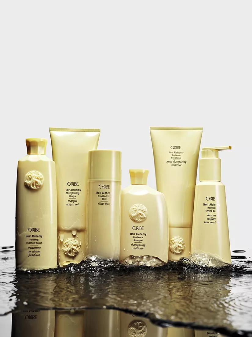 Oribe Hair Alchemy shampoo, conditioner and styling products in gold bottles, displayed with water splash studio shot