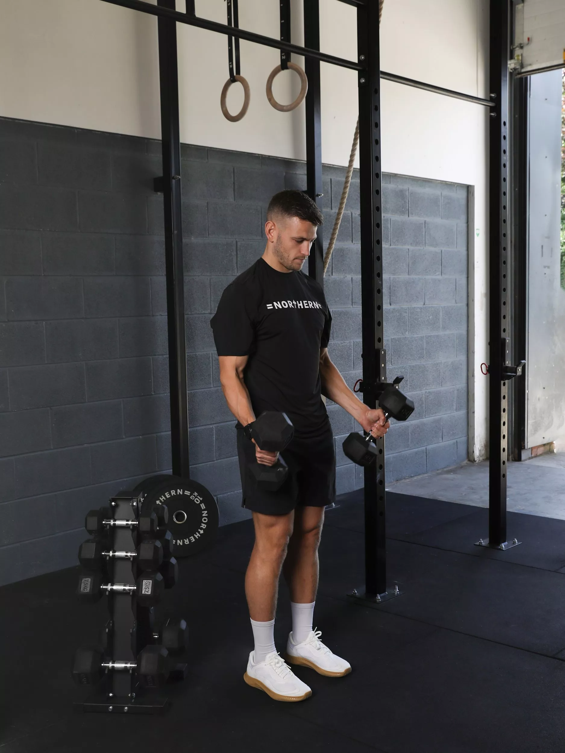Man performing dumbbell bicep curls in a modern CrossFit gym, with rings and weight rack in background