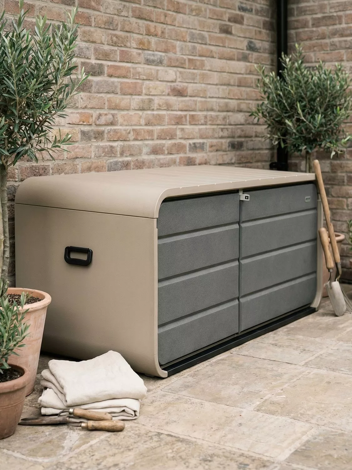 Weatherproof outdoor garden storage box beside brick wall, with patio tools and potted olive trees on stone paving