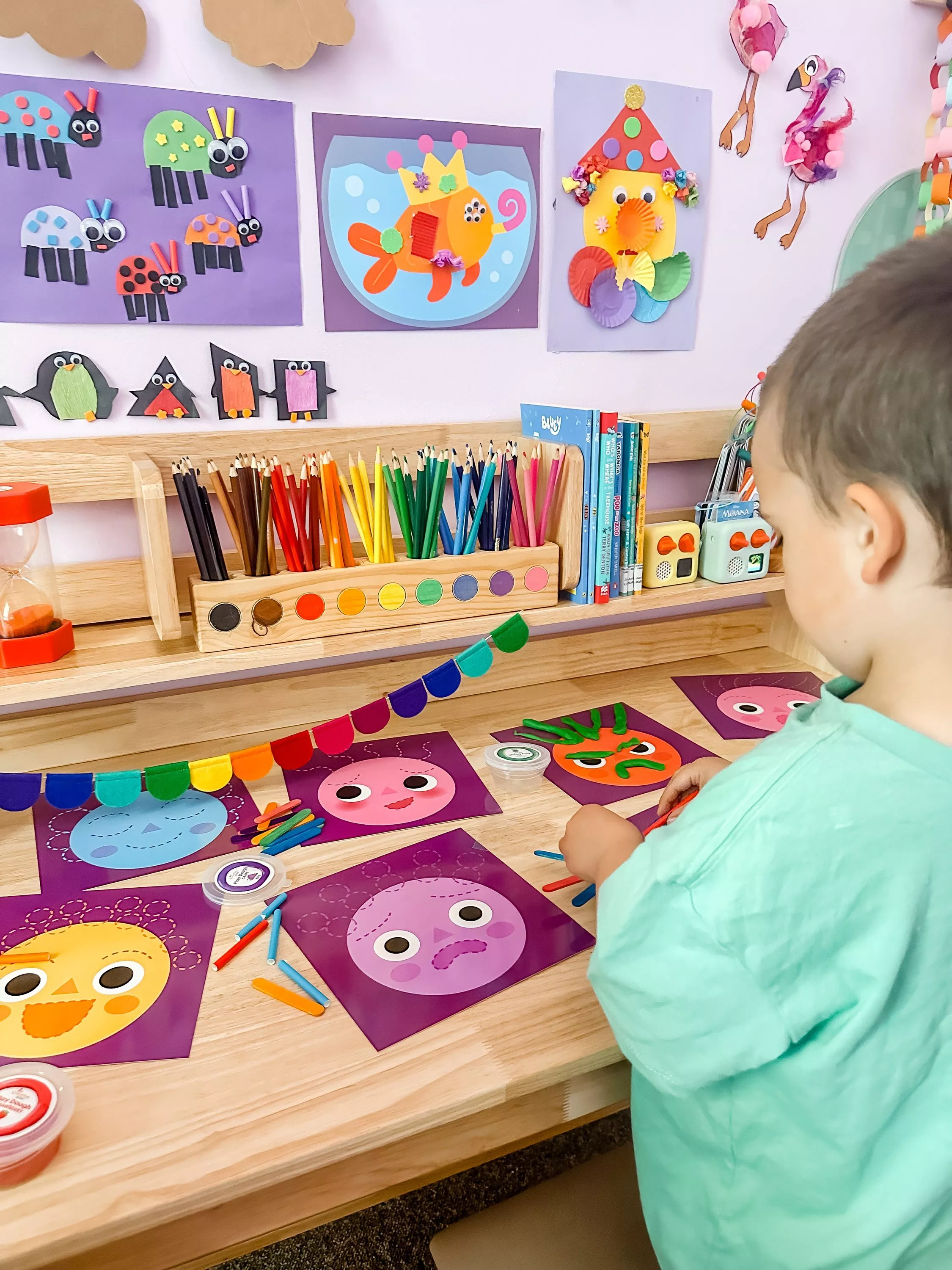 Child doing colourful preschool craft activity with paper faces, pencils and art display on classroom wall