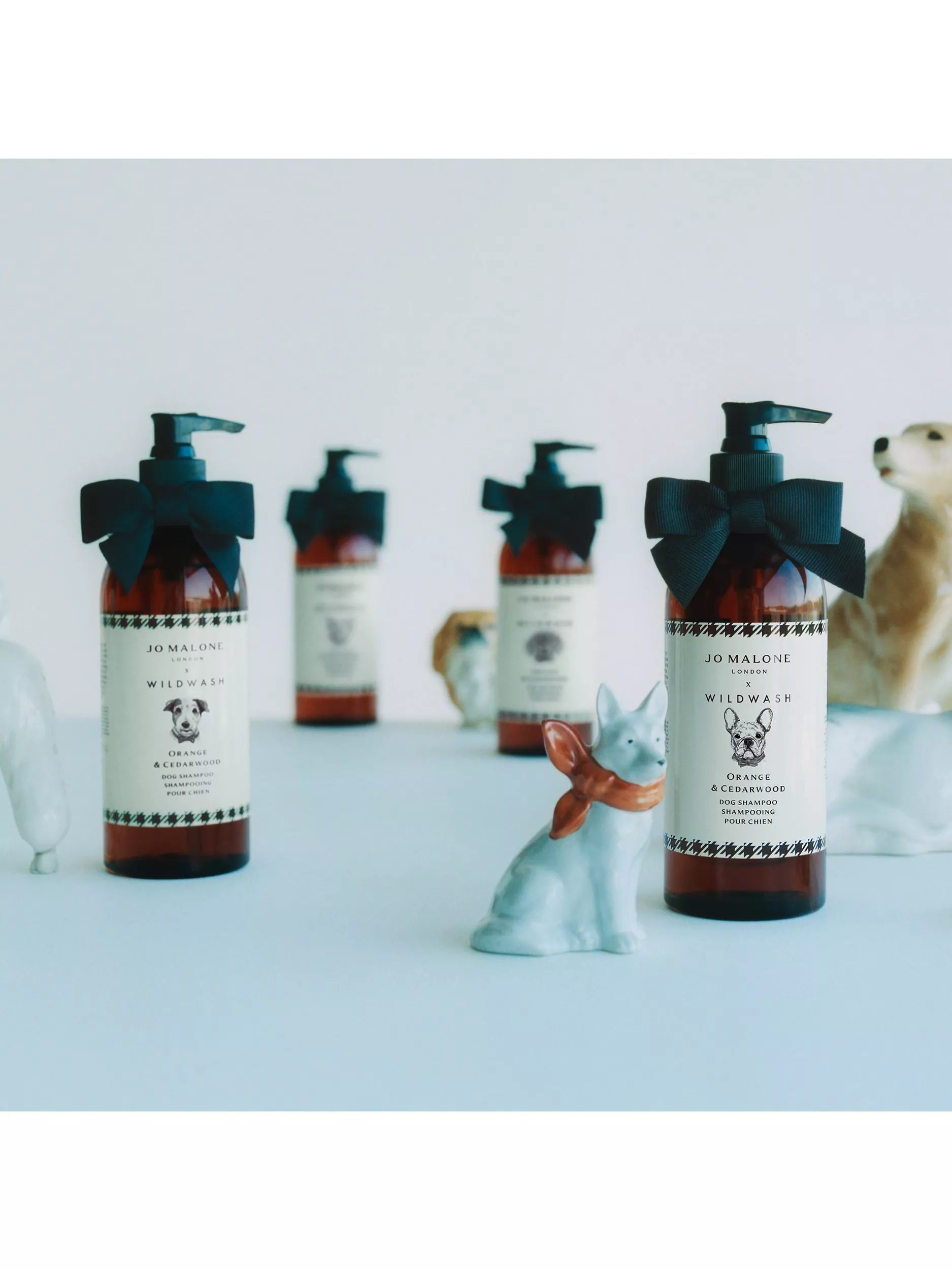 Jo Malone London Wildwash dog shampoo bottles with black bows, styled with ceramic dog figurines on a pale backdrop