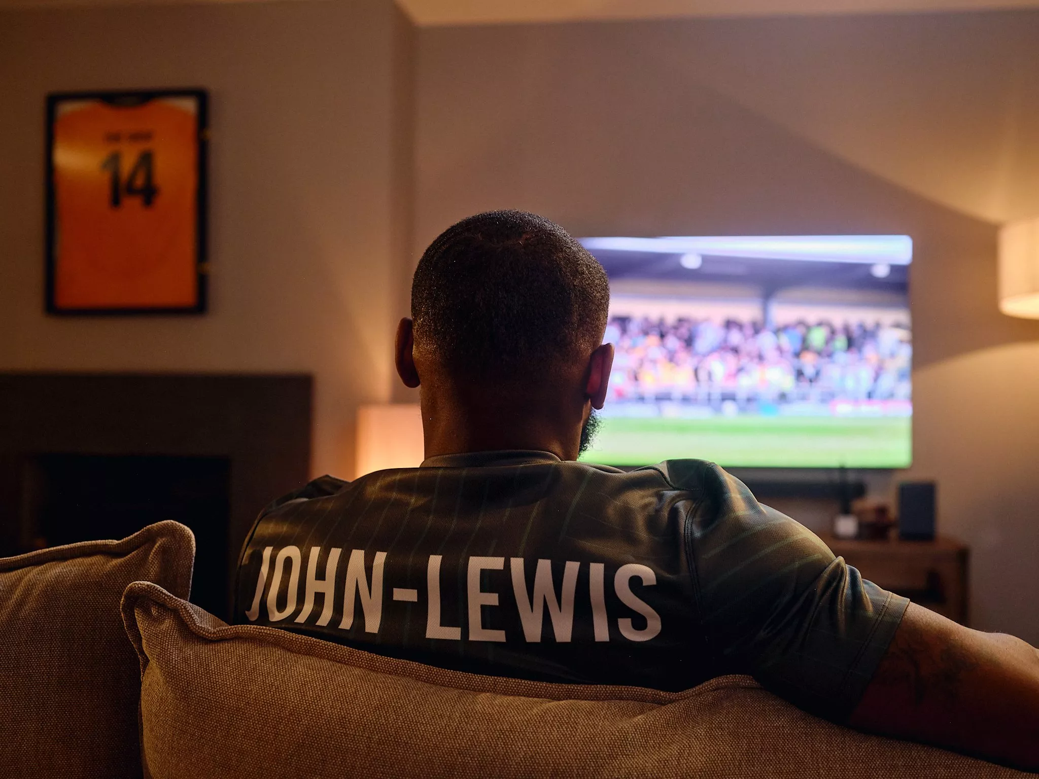 Man wearing John-Lewis football shirt watching a live match on TV in a cosy living room at night