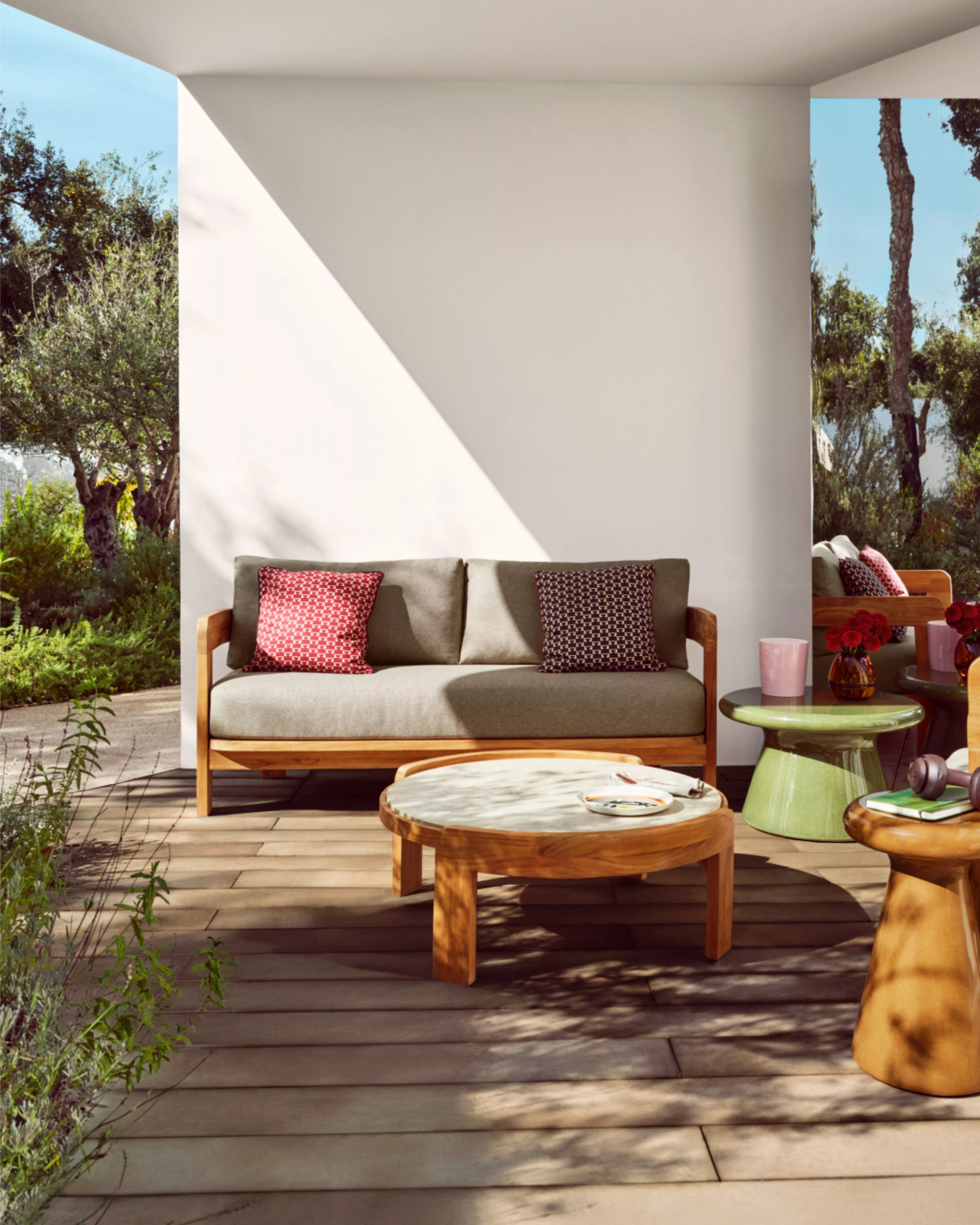 Modern outdoor seating area with wooden furniture, colourful cushions, and tables. Trees and greenery surround the space.
