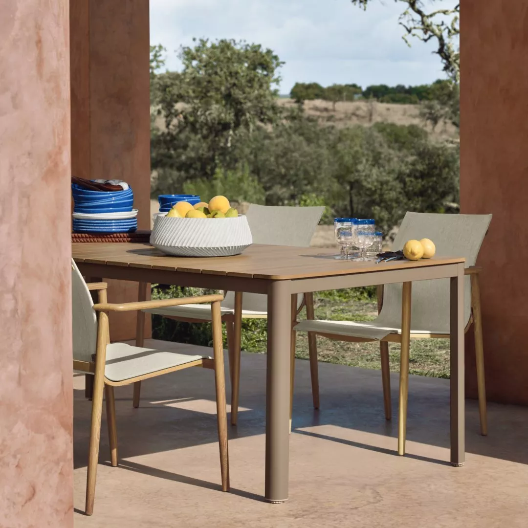 Modern outdoor dining table and chairs on patio with fruit bowl, glasses and countryside view in sunlight