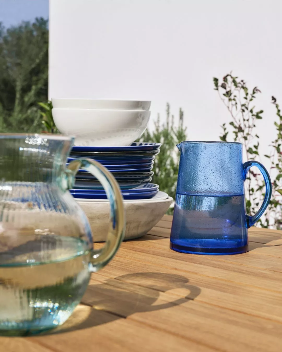 Outdoor table setting with stacked blue plates, white bowls and blue glass water jug in sunny garden patio scene