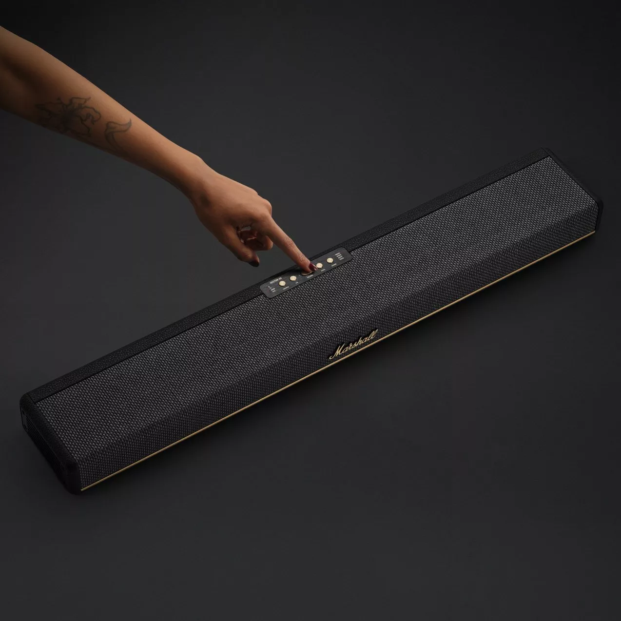 A hand adjusts settings on a sleek black soundbar with mesh design and gold accents on a dark background.
