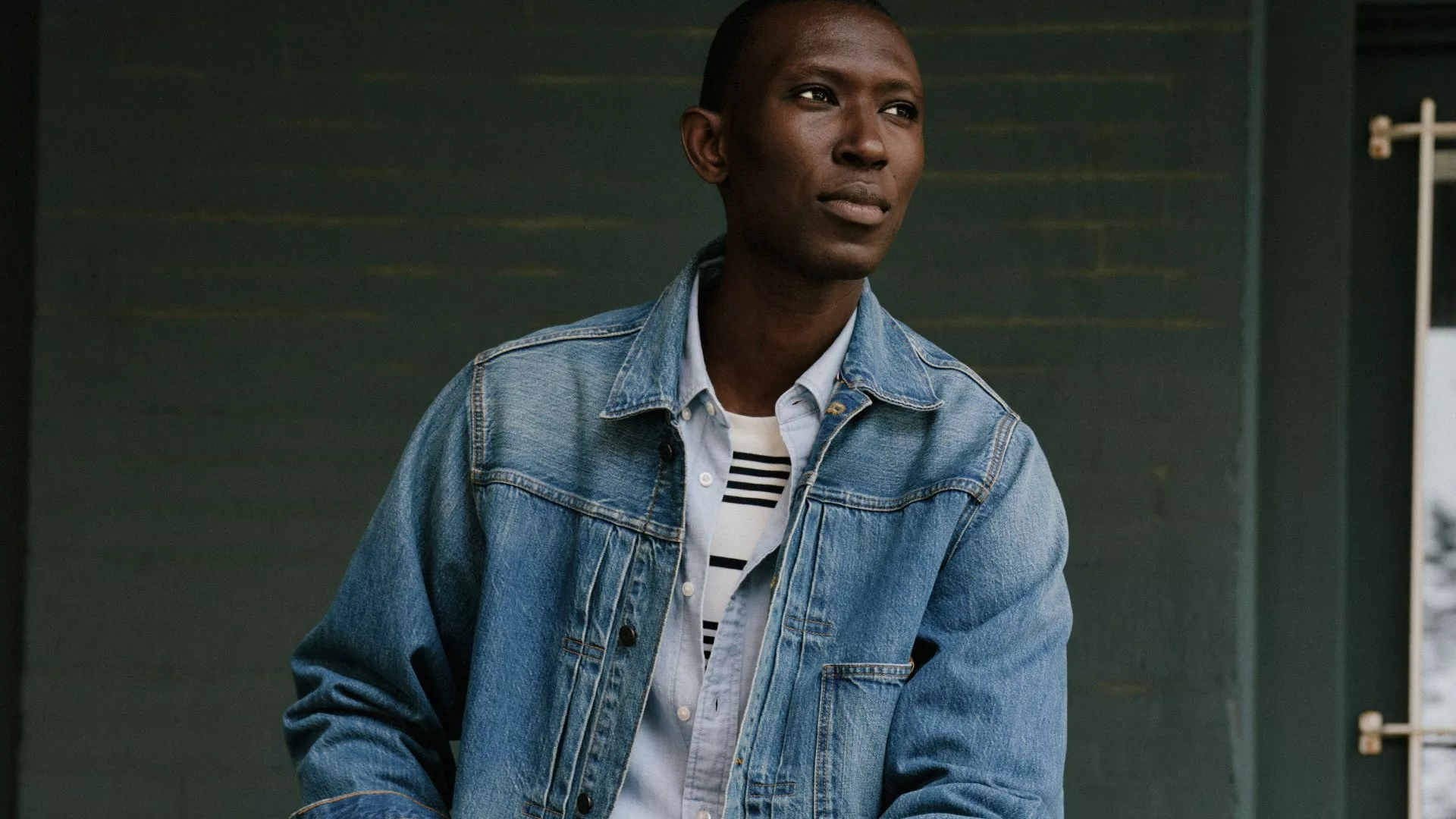 A man wearing a striped shirt, a light blue button-down, and a classic denim jacket against a dark green brick wall.