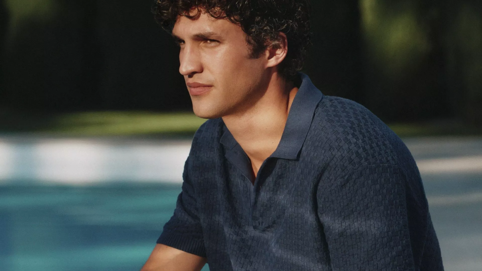 Close-up portrait of a man in a navy polo shirt by a swimming pool, looking thoughtful in sunlight