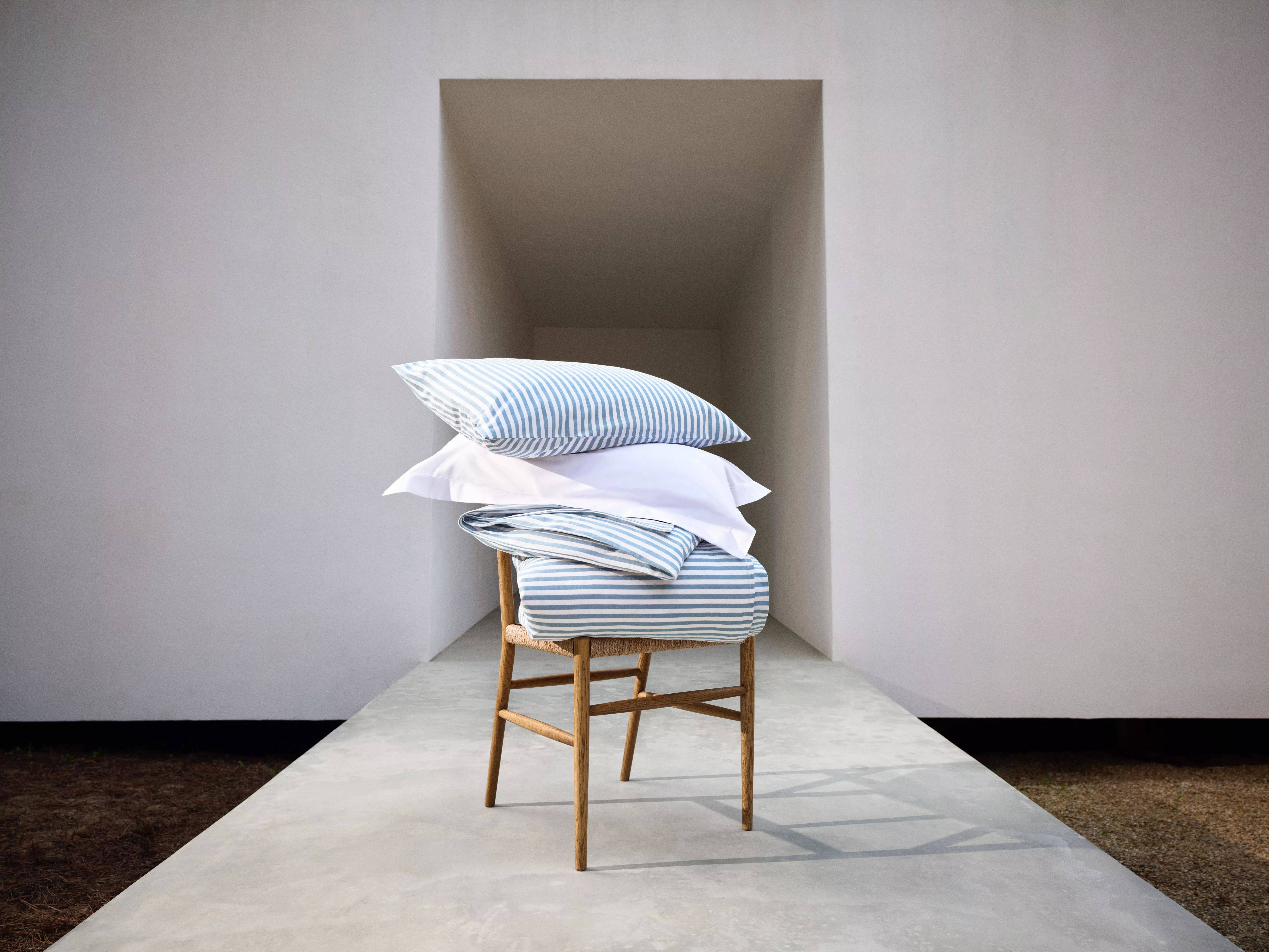 Blue and white striped bedding set with pillows stacked on a wooden chair in a minimalist hallway setting