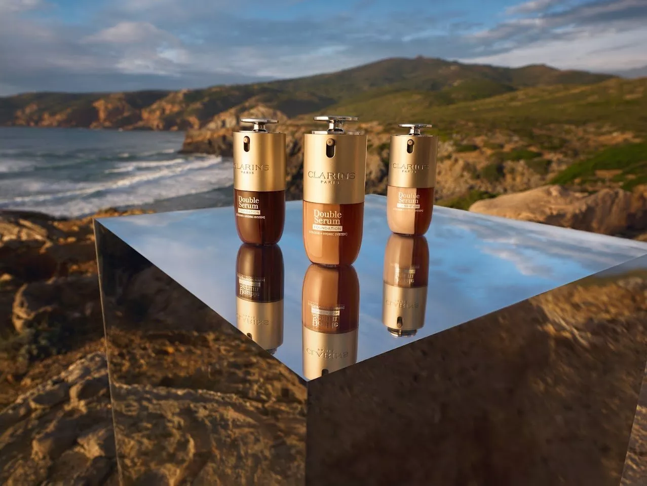 Three Clarins Double Serum bottles on a mirrored cube by the coast, reflecting the scenic ocean and hills.