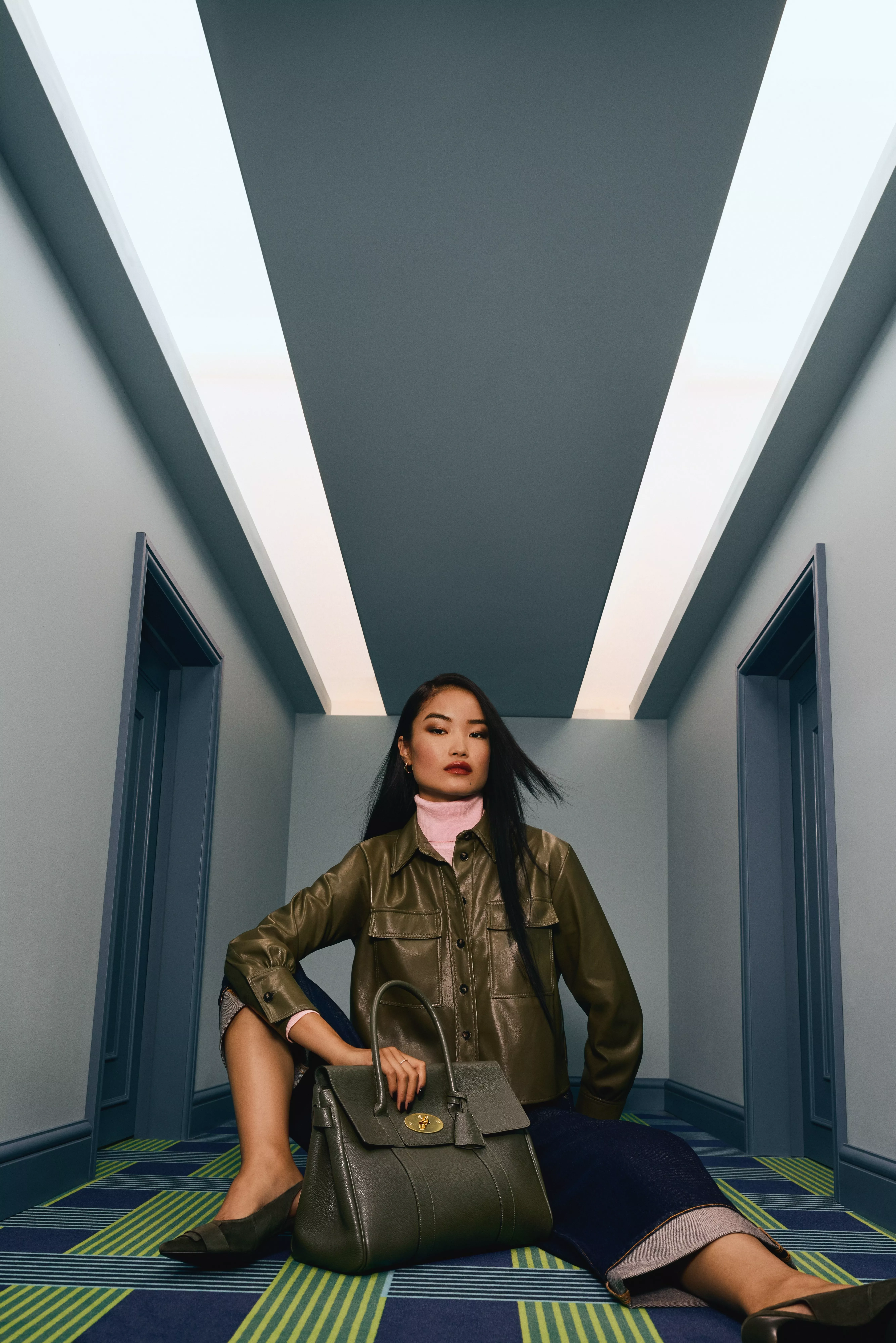 Woman in green leather jacket sits on the floor with a Mulberry leather handbag in front of her