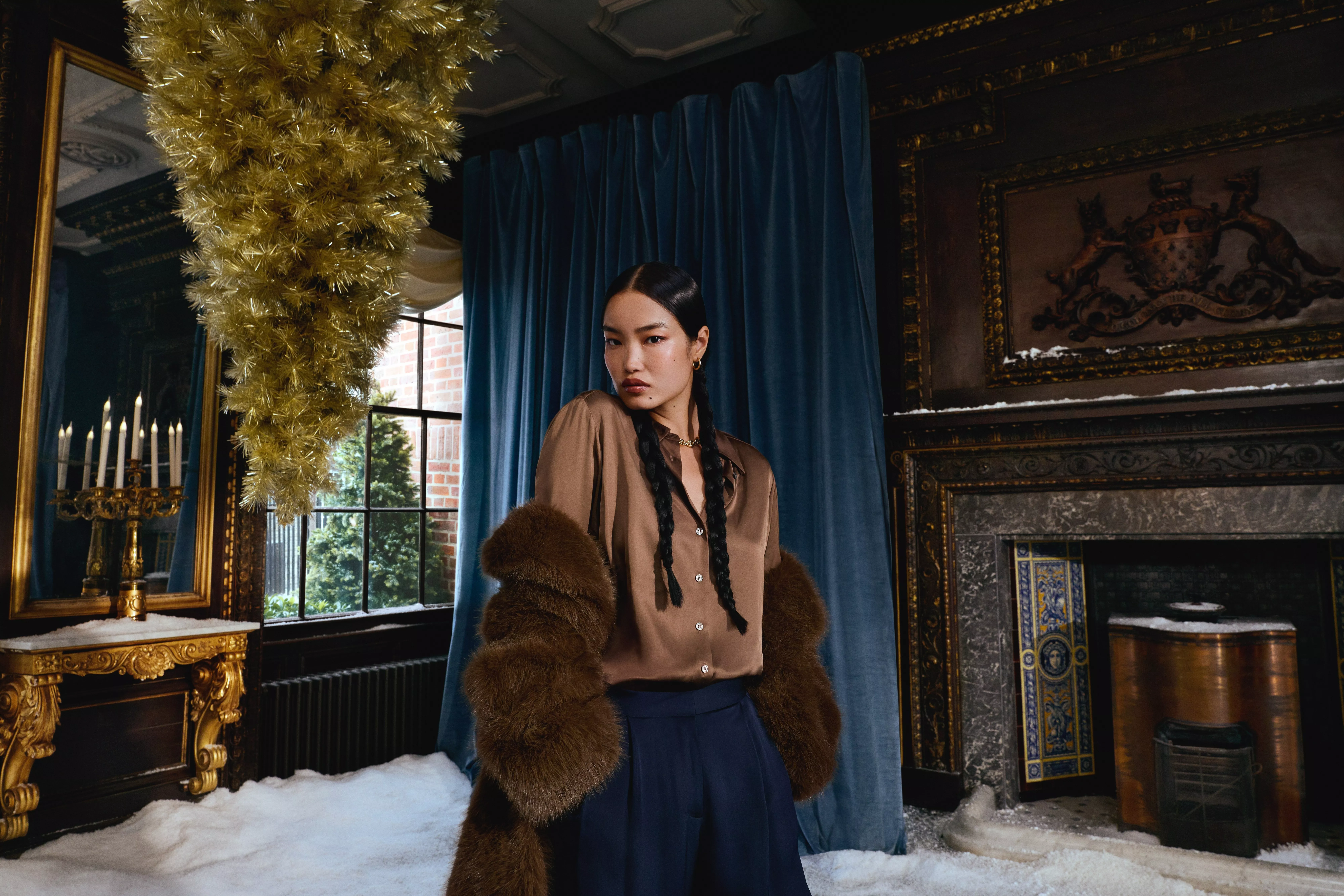 A person with long, dark braided hair looks toward the camera. They are wearing a brown silk button-up shirt, a dark navy skirt, and a voluminous brown faux-fur coat draped over their shoulders.