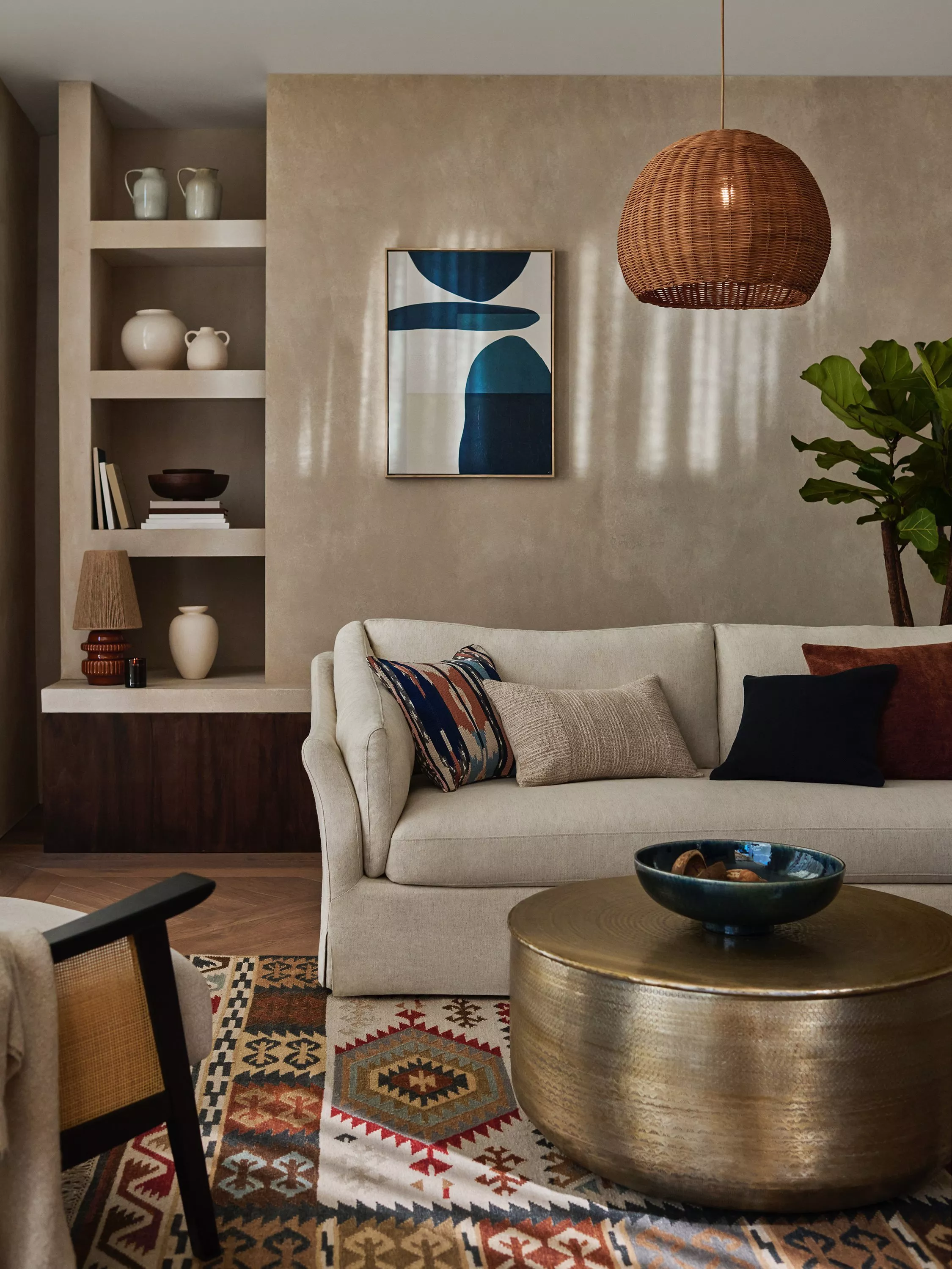 A neutral styled living room with a beige sofa, round bronze coffee table, ornate rug, rattan lampshade and abstract picture on the wall next to shelving with objects displayed