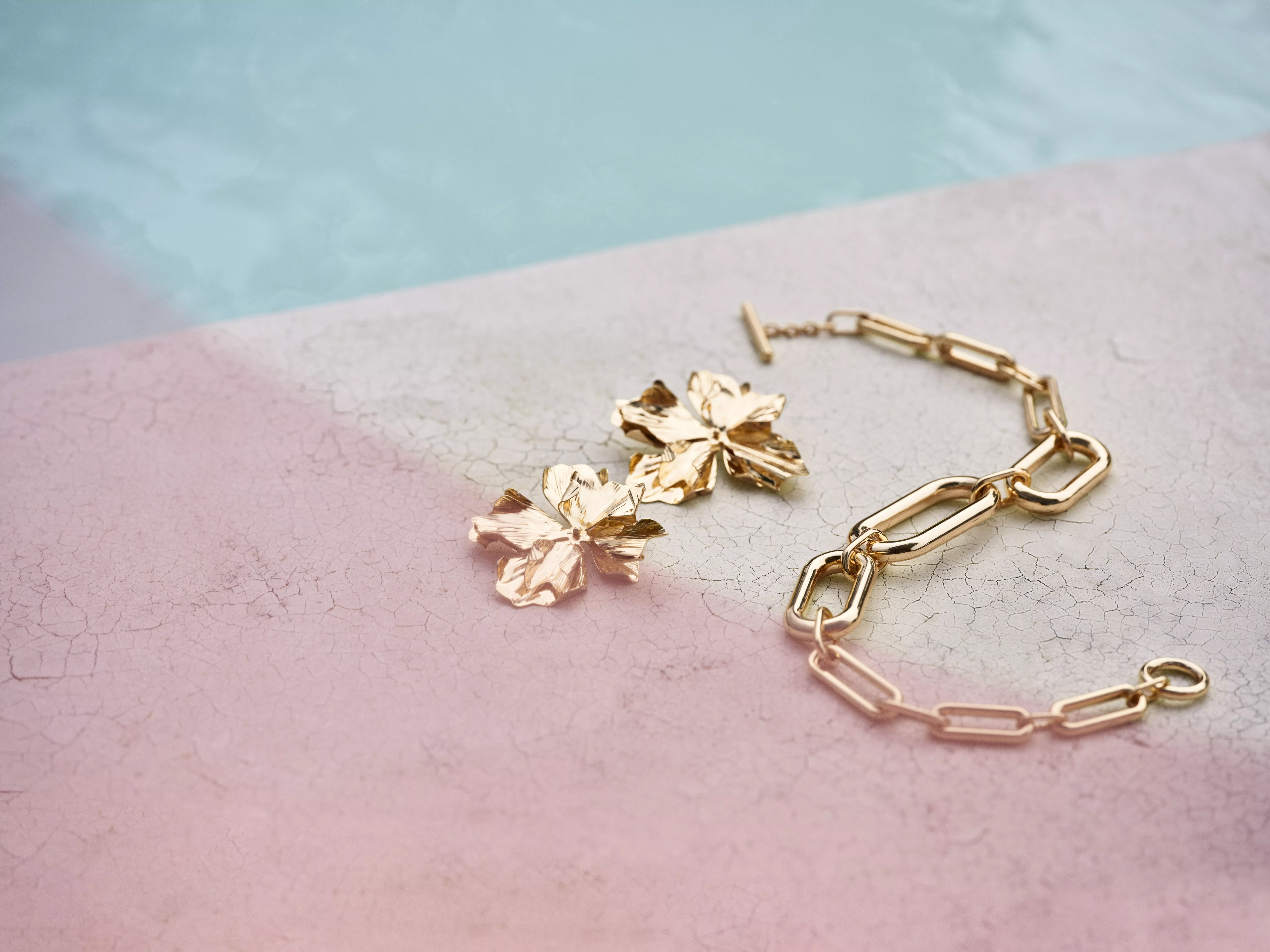 Gold chain bracelet and floral earrings on a pink textured surface by a pool, creating a stylish, elegant vibe.