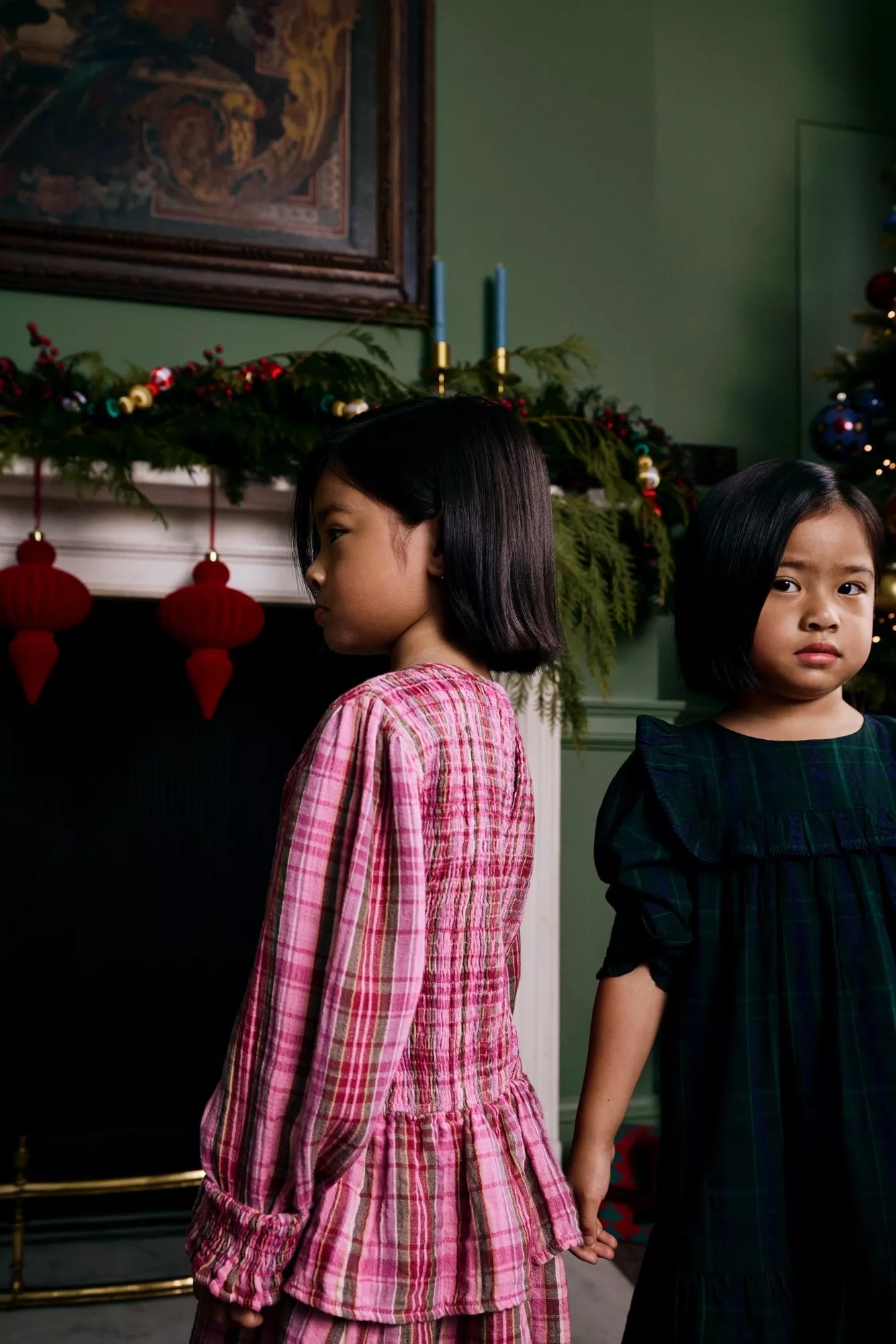 Two young girls in festive plaid dresses stand in front of a Christmas decorated fireplace
