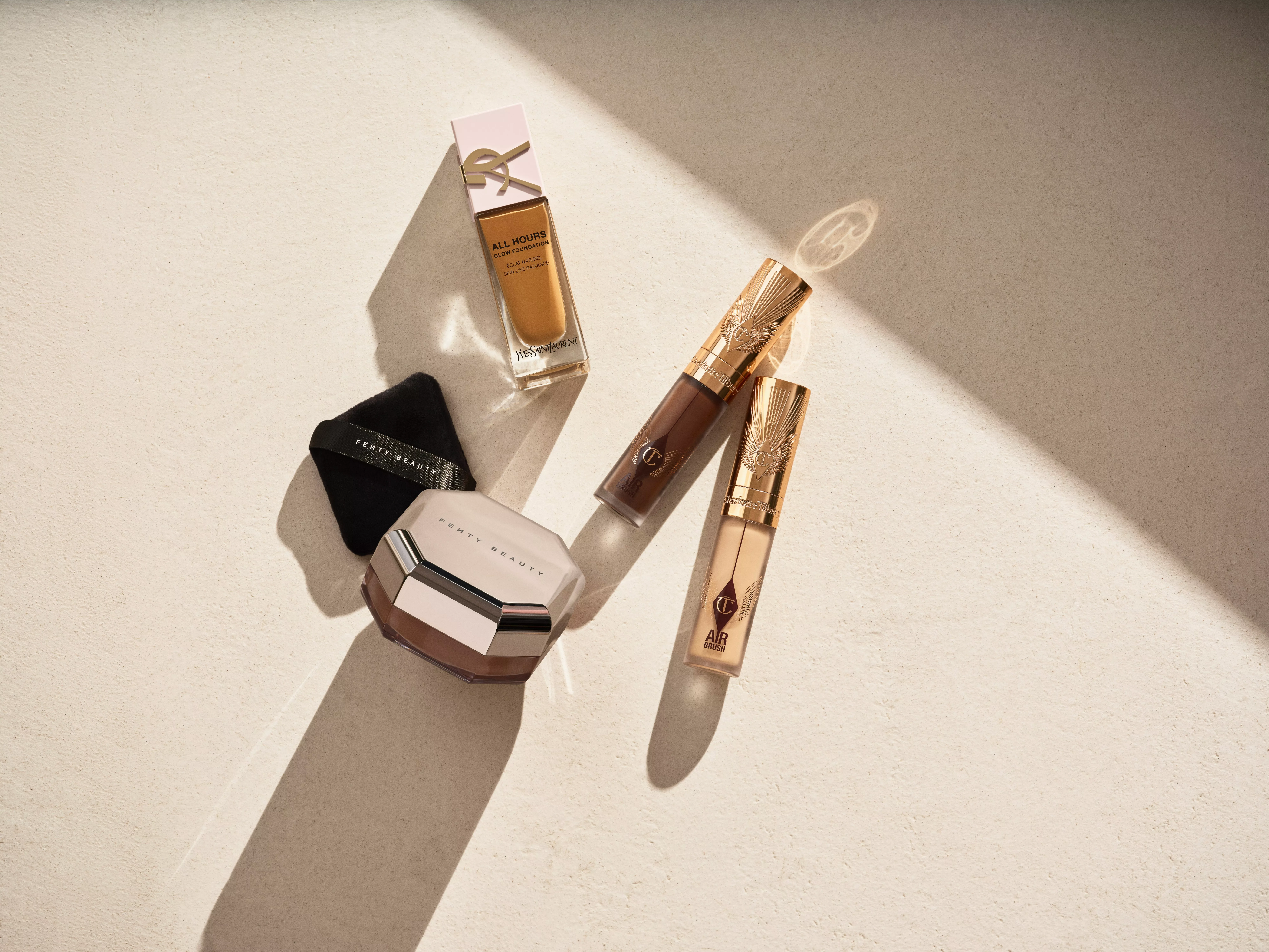 Luxury makeup products flat lay: foundation, concealer and Fenty Beauty compact in warm sunlight on beige surface