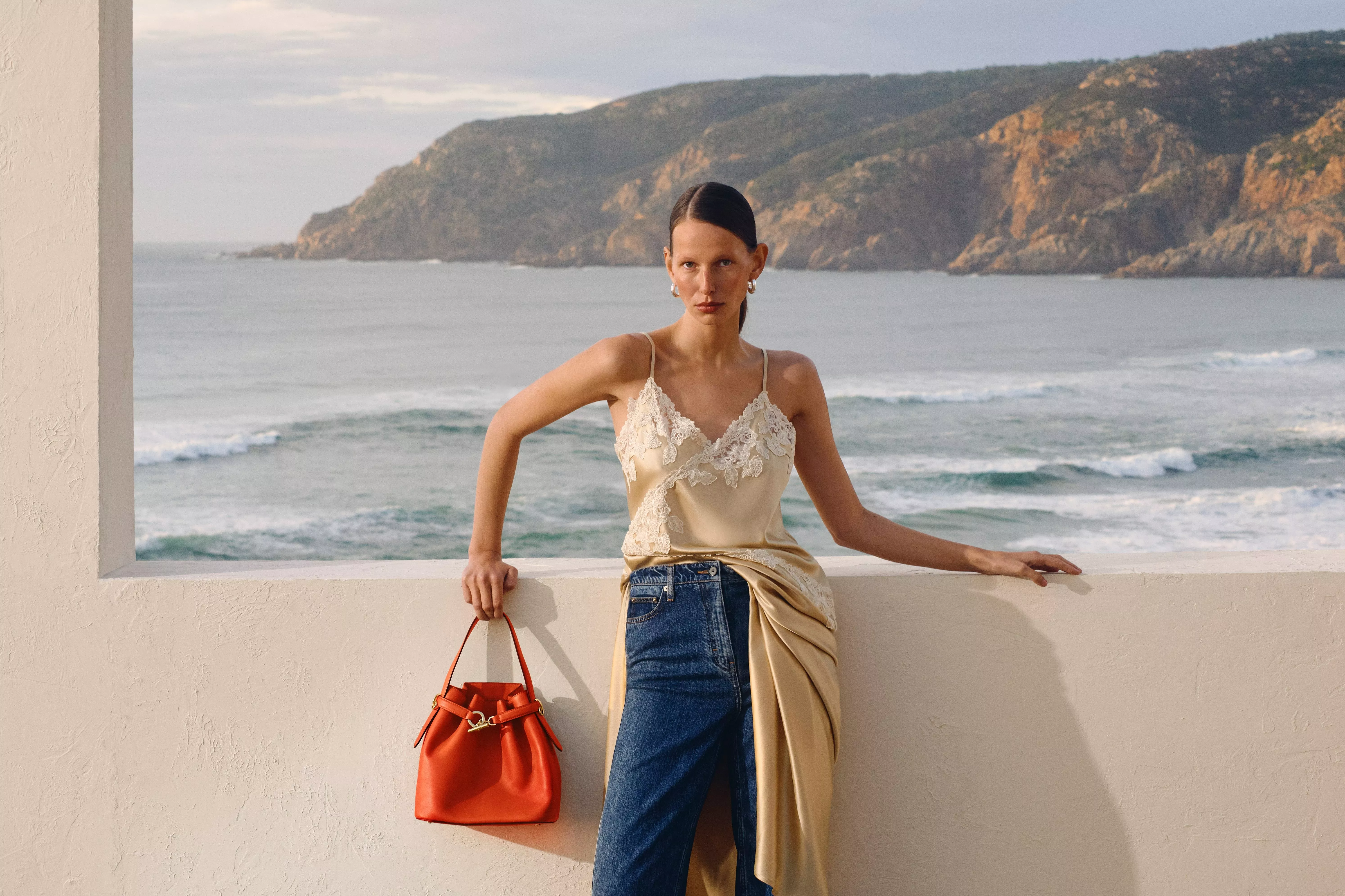 Model in a lace dress and jeans holding an orange handbag stood in front of a beach landscape