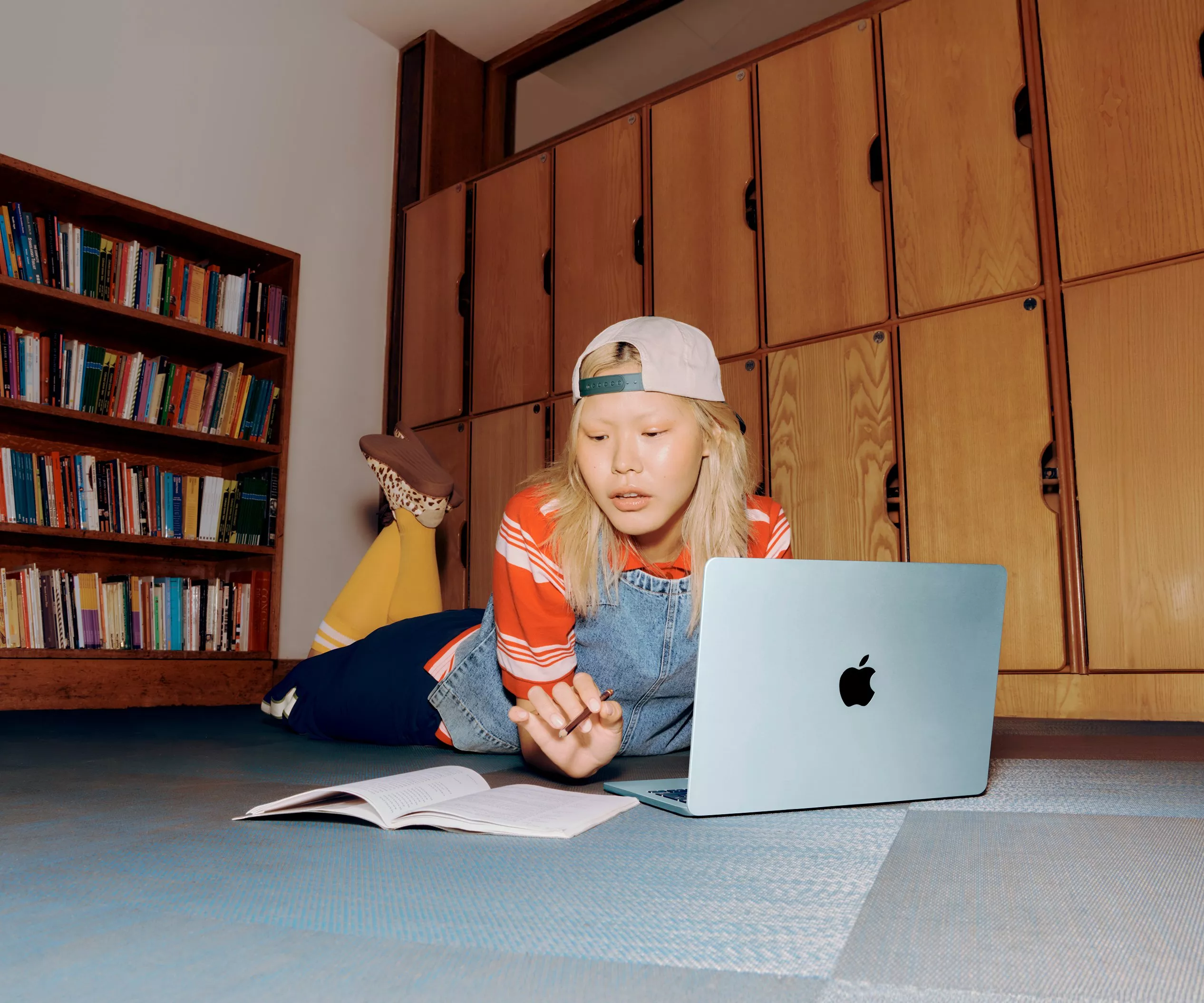 Student studying on laptop on floor with notebook in library room, wearing cap and casual clothes