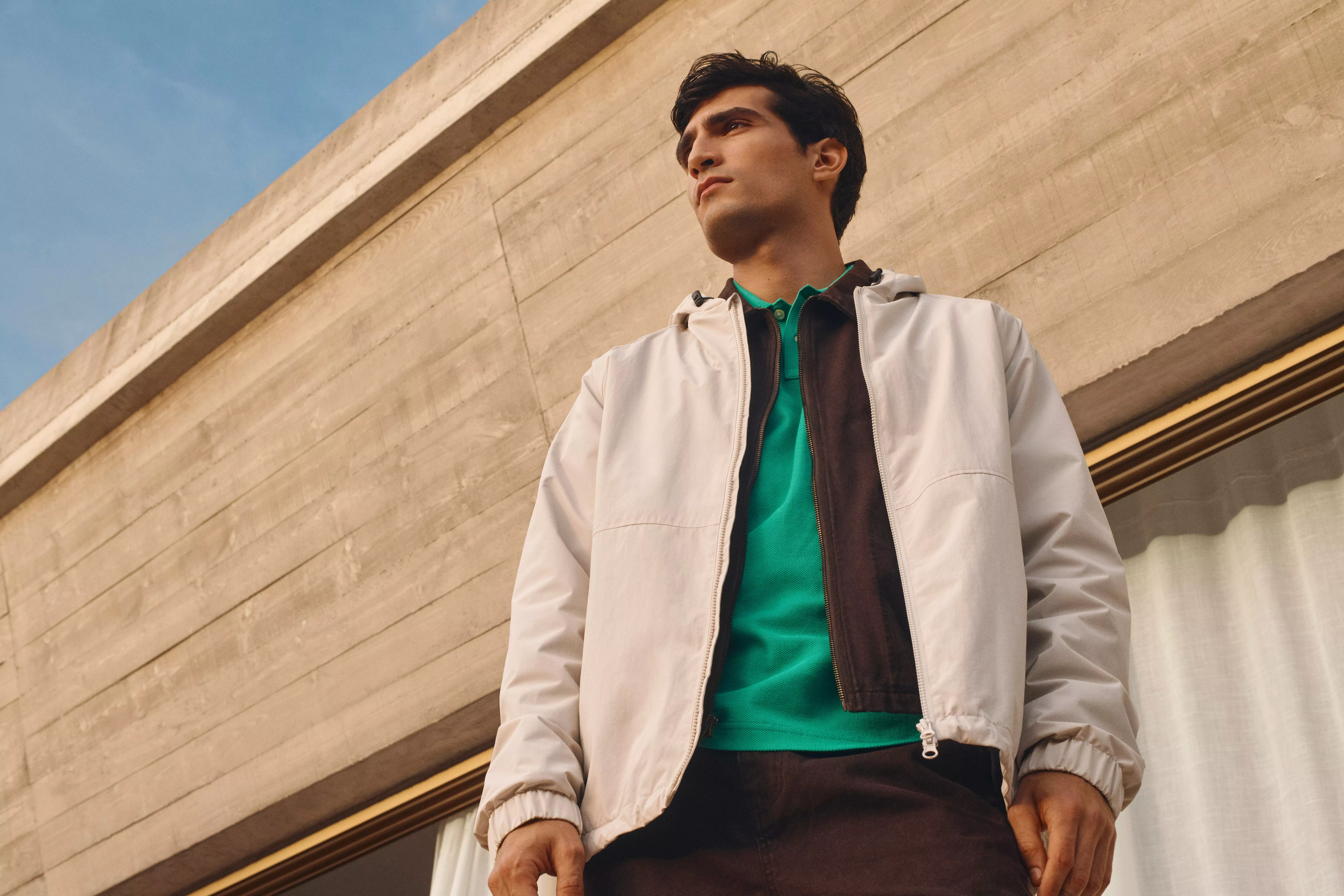 A person in a white jacket and green shirt stands confidently against a modern concrete building backdrop.