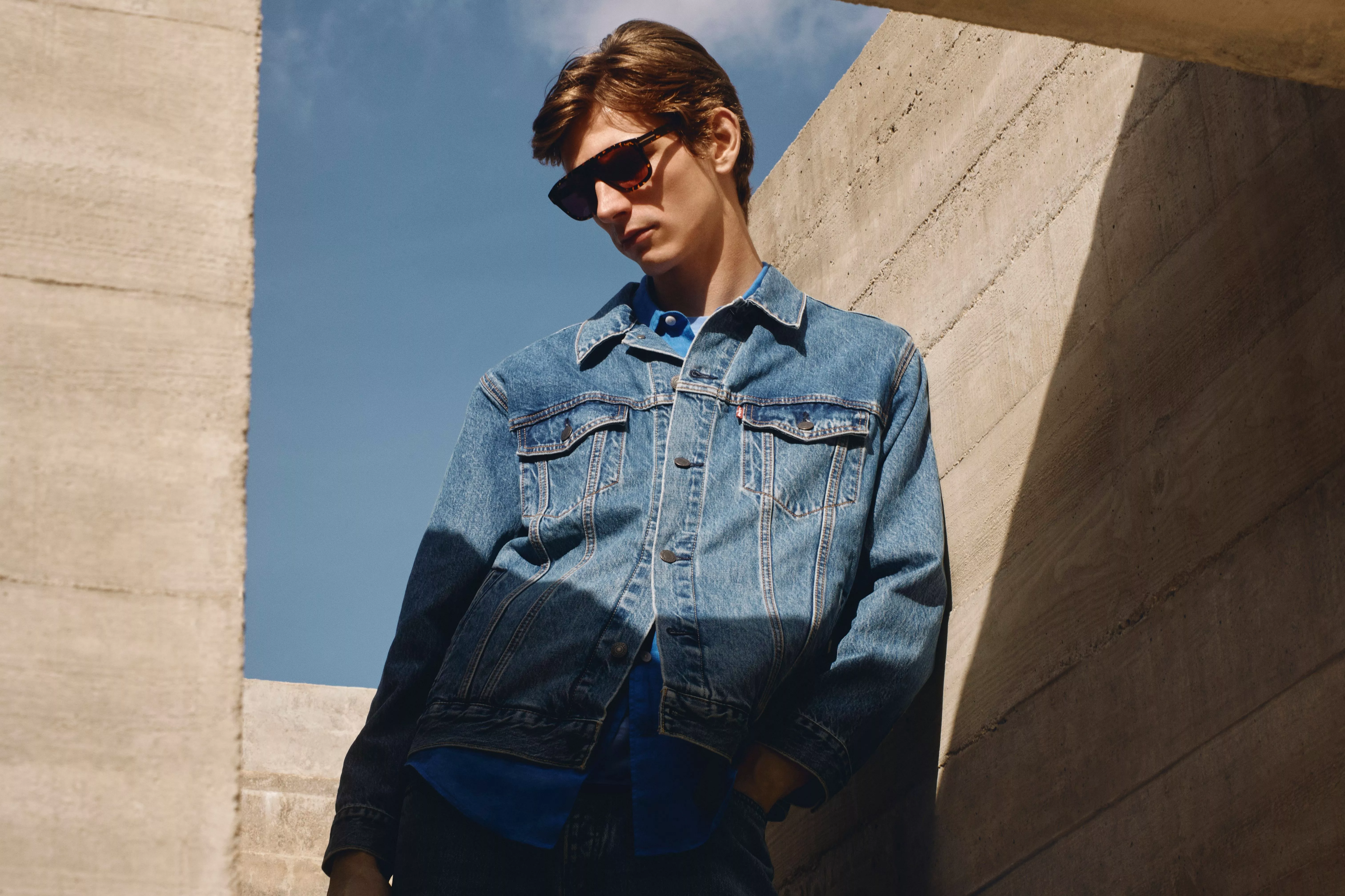 A low-angle shot of a male model wearing a classic blue denim jacket and dark sunglasses.