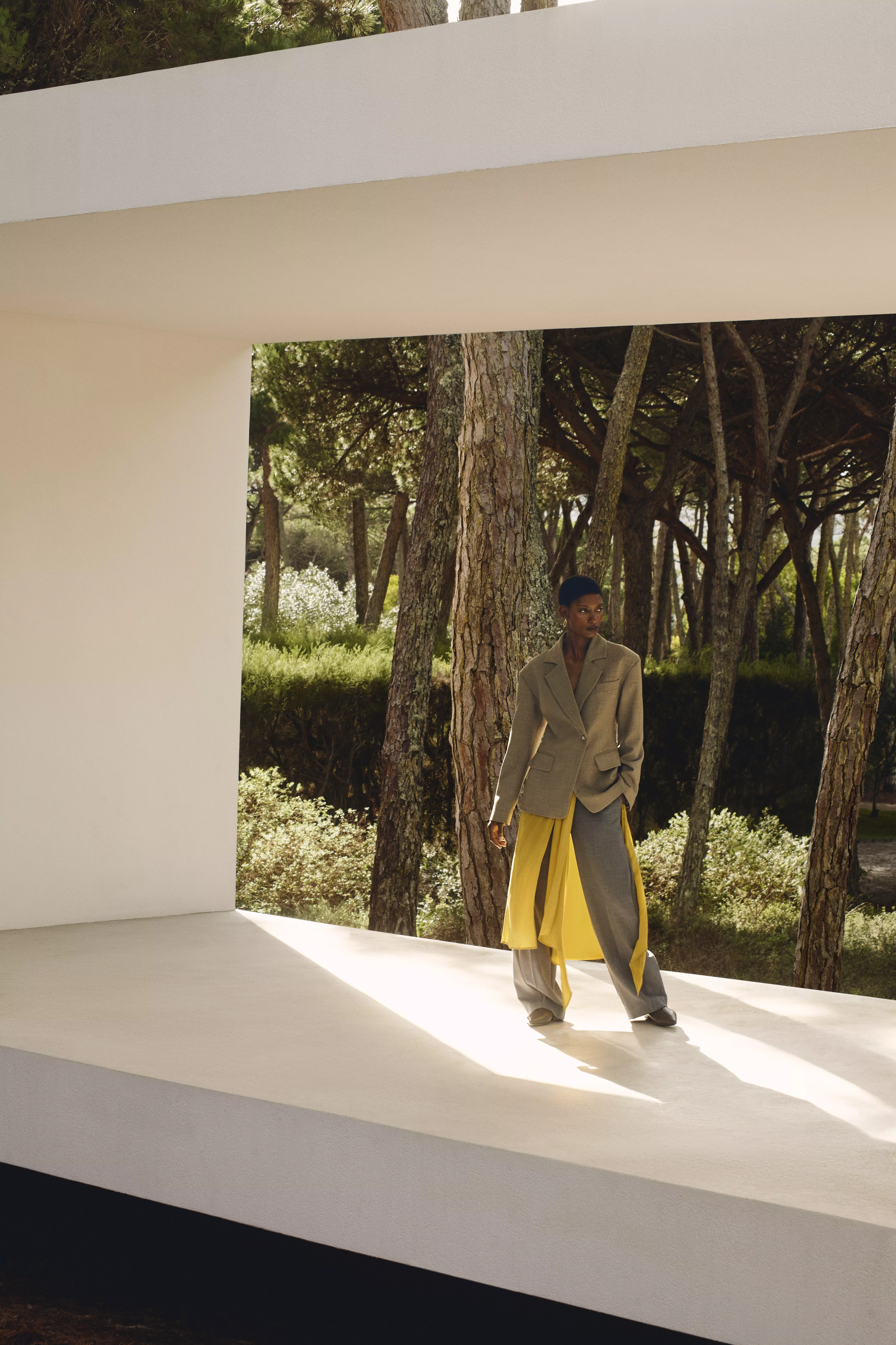 A woman in a stylish taupe trouser suit with a long yellow blouse, accessorised with gold earrings