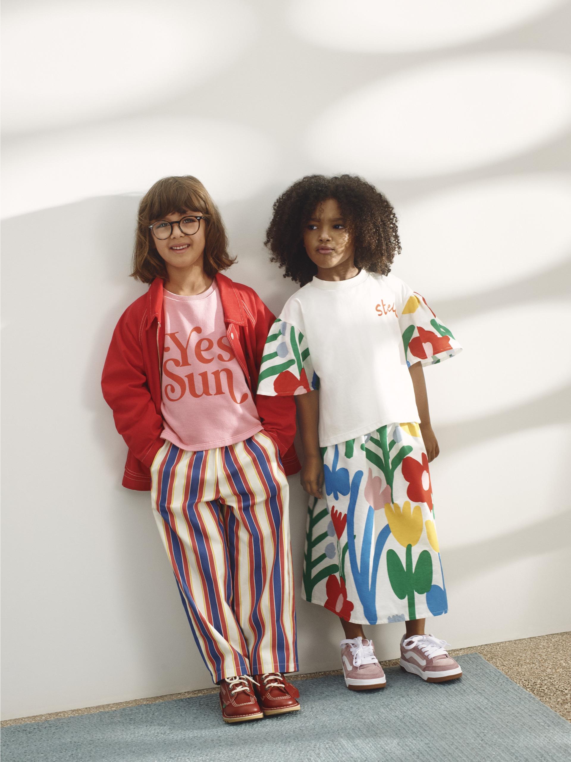 Two children modelling colourful kids’ outfits, striped trousers and floral skirt, standing against a sunlit wall