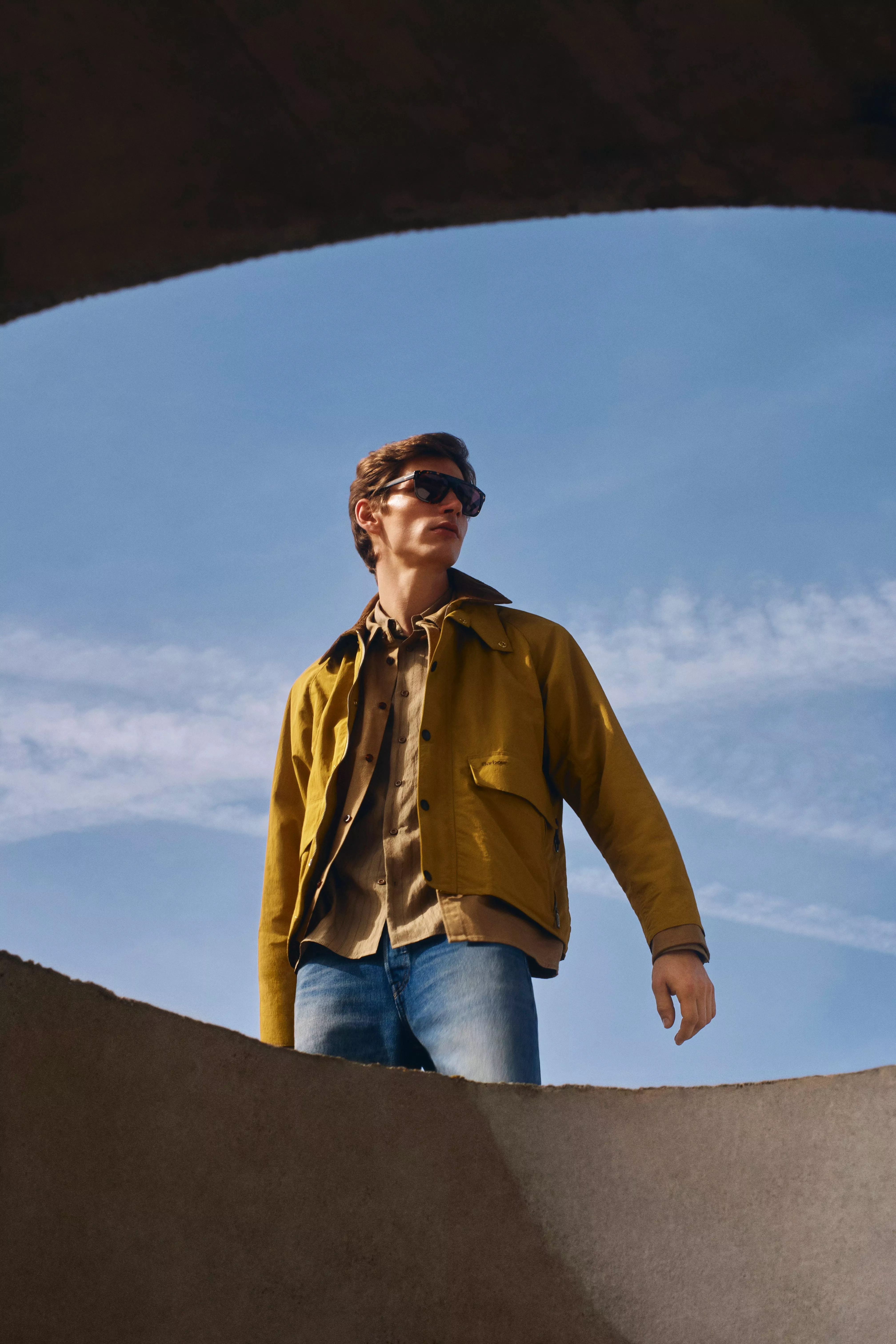 Man in yellow jacket and sunglasses stands against a clear blue sky, looking to the side confidently.