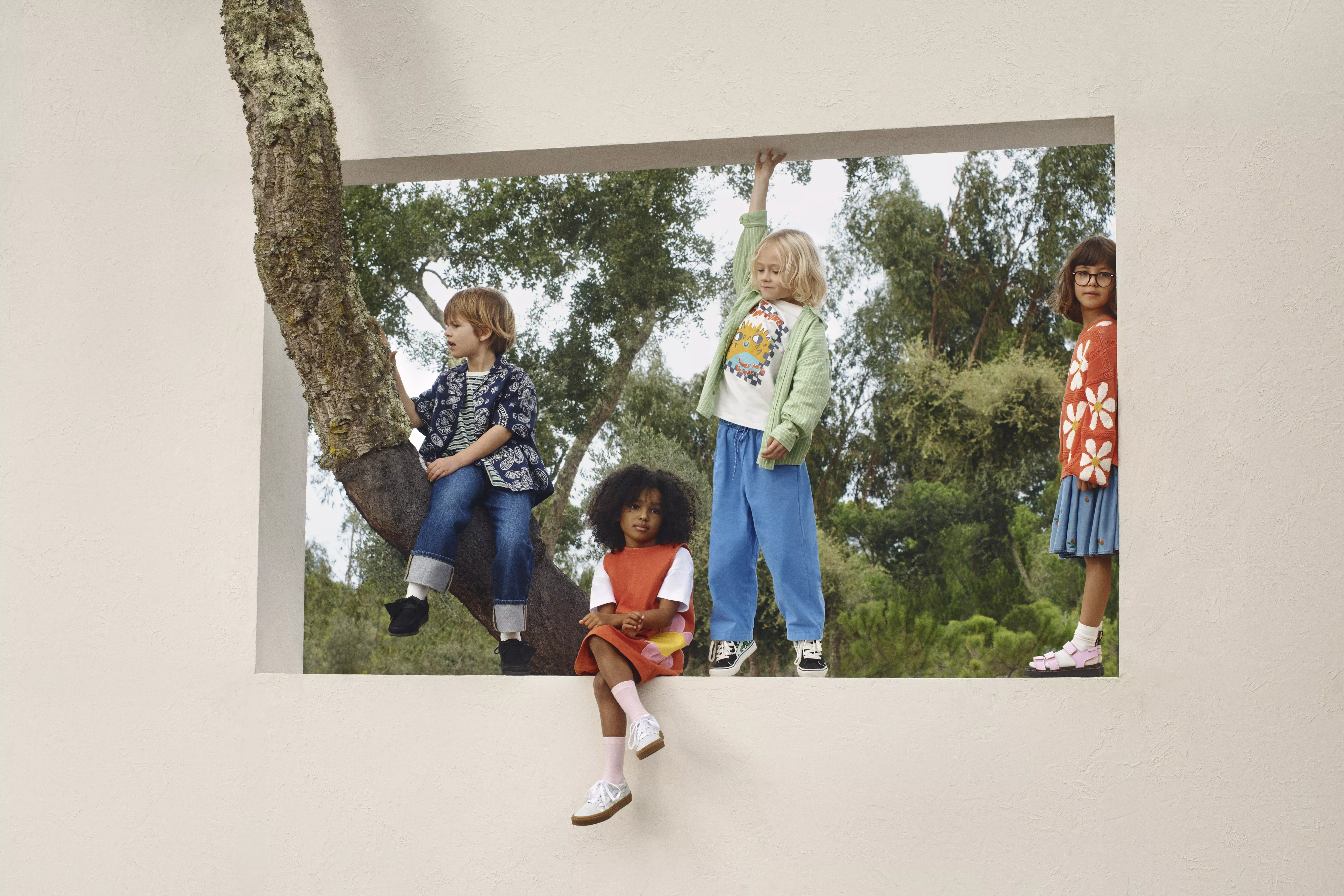 Children wearing colourful outfits play on a tree through a rectangular wall opening, surrounded by greenery.