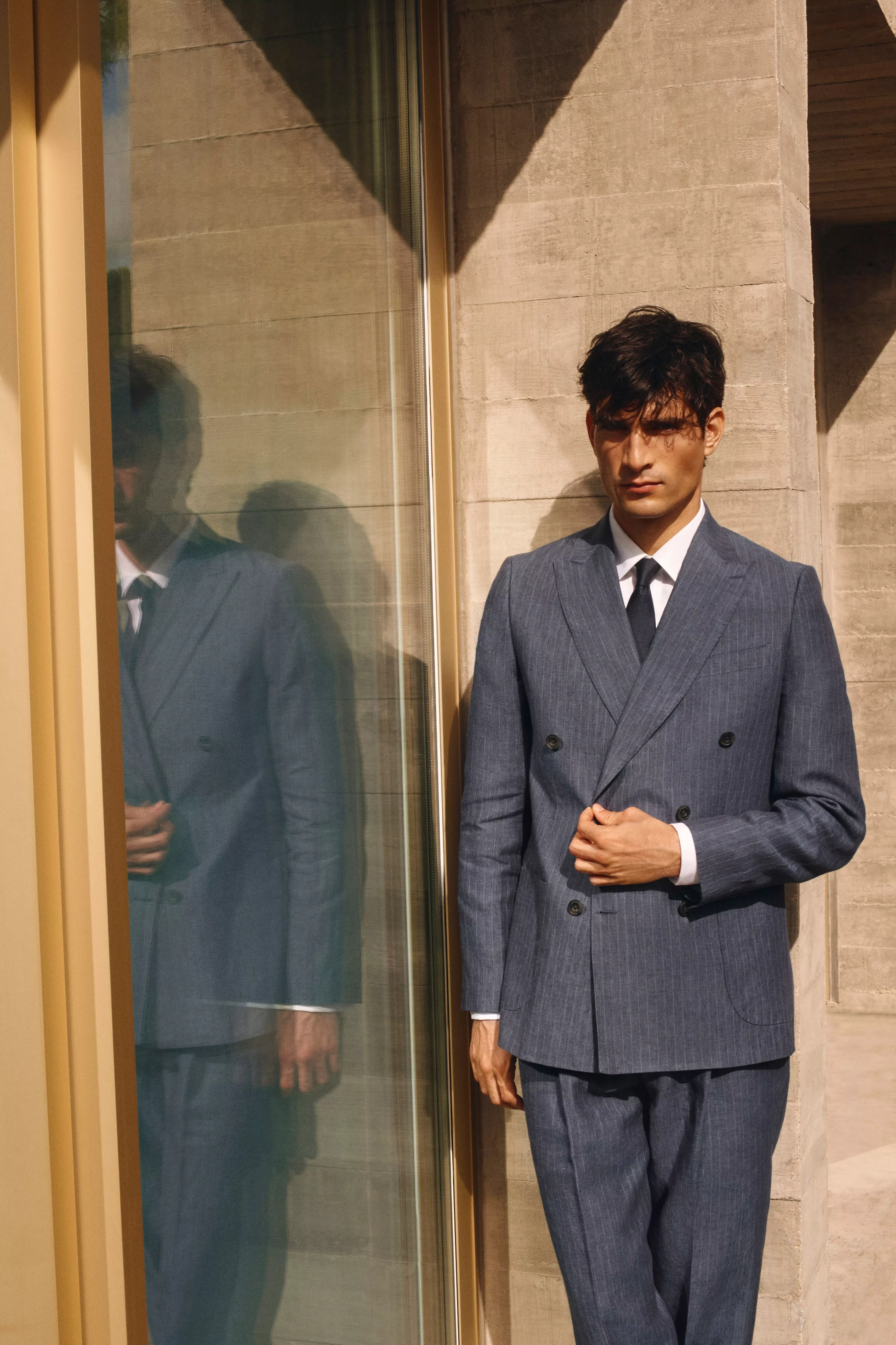 Man in a blue double-breasted suit stands against a concrete wall, with his reflection visible in the glass.
