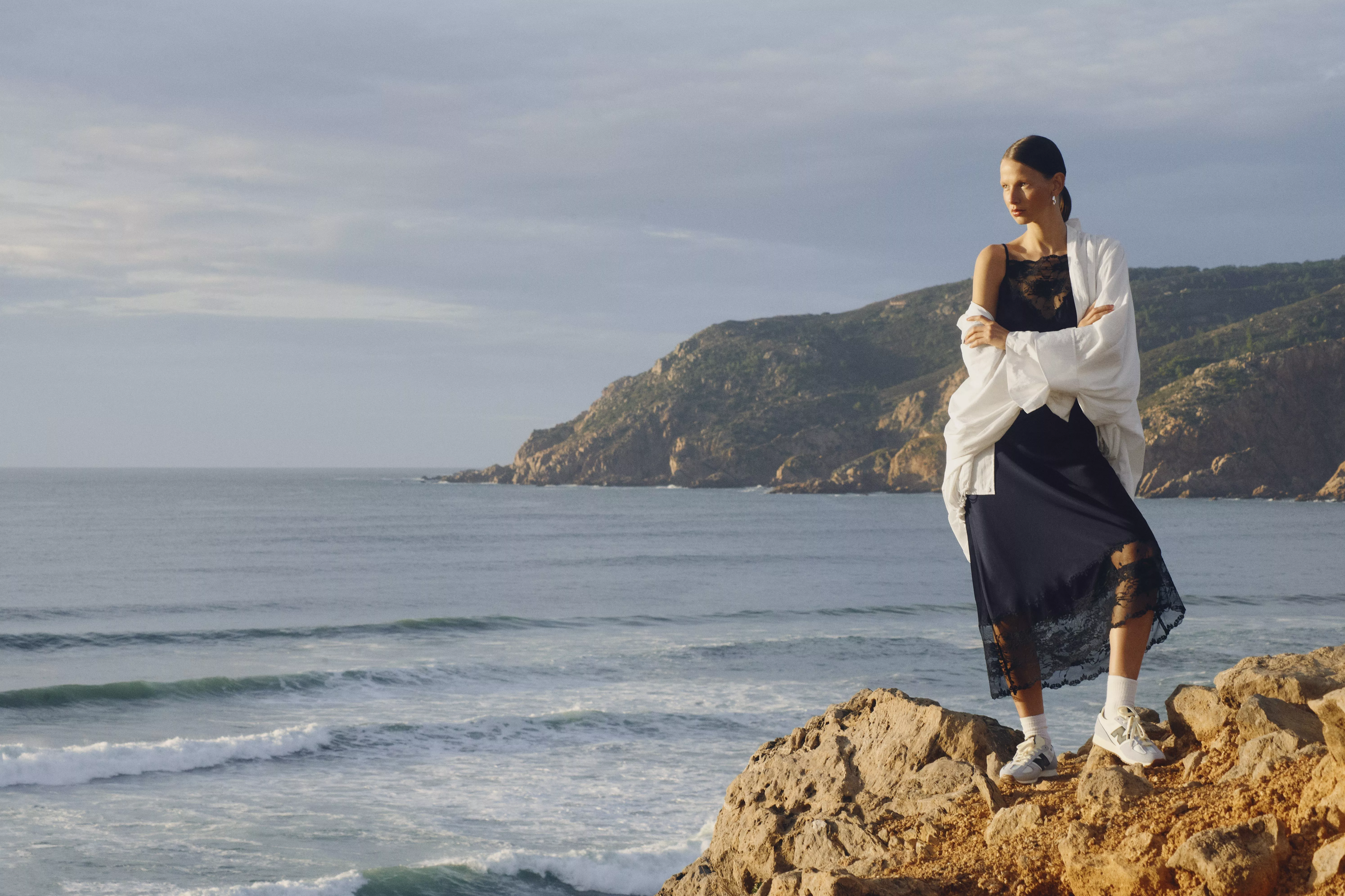 woman wearing black lace maxi dress and white cover up and trainers standing on a rock on the coast