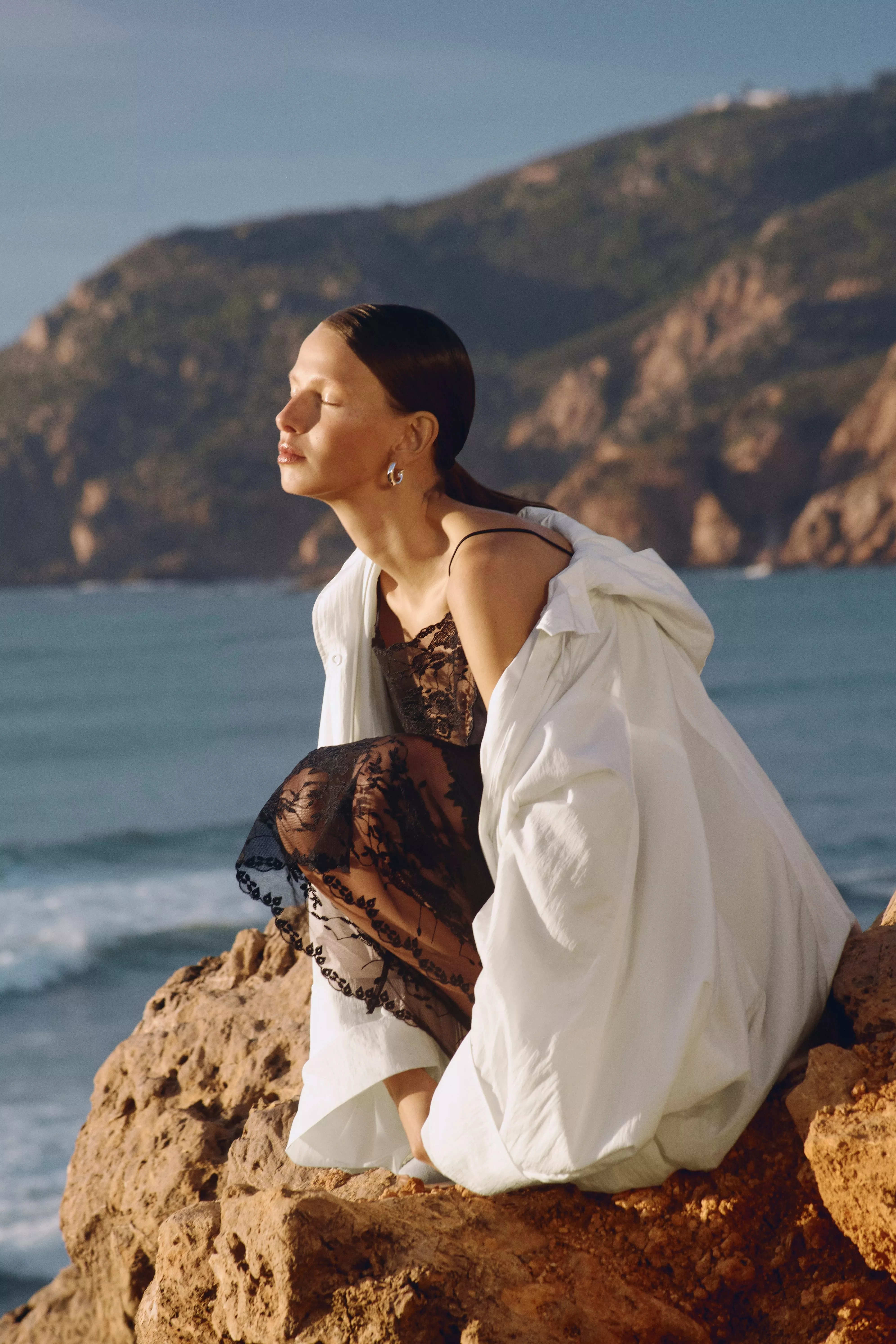 Fashion model in black lace dress and white shirt on rocky beach, coastal cliffs and sea backdrop at sunset