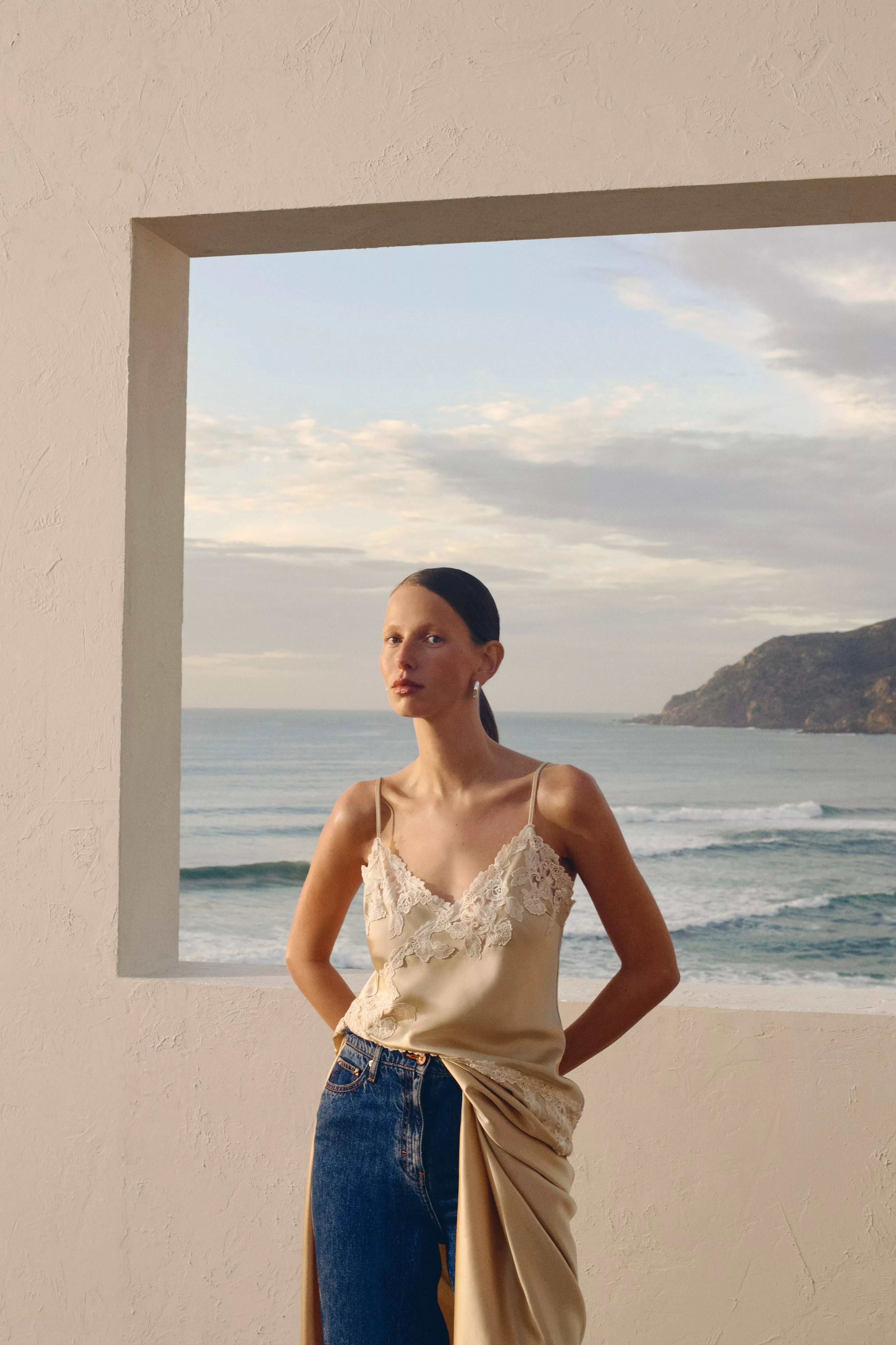A woman in a lace-trimmed camisole and jeans poses against a coastal backdrop framed by a concrete window