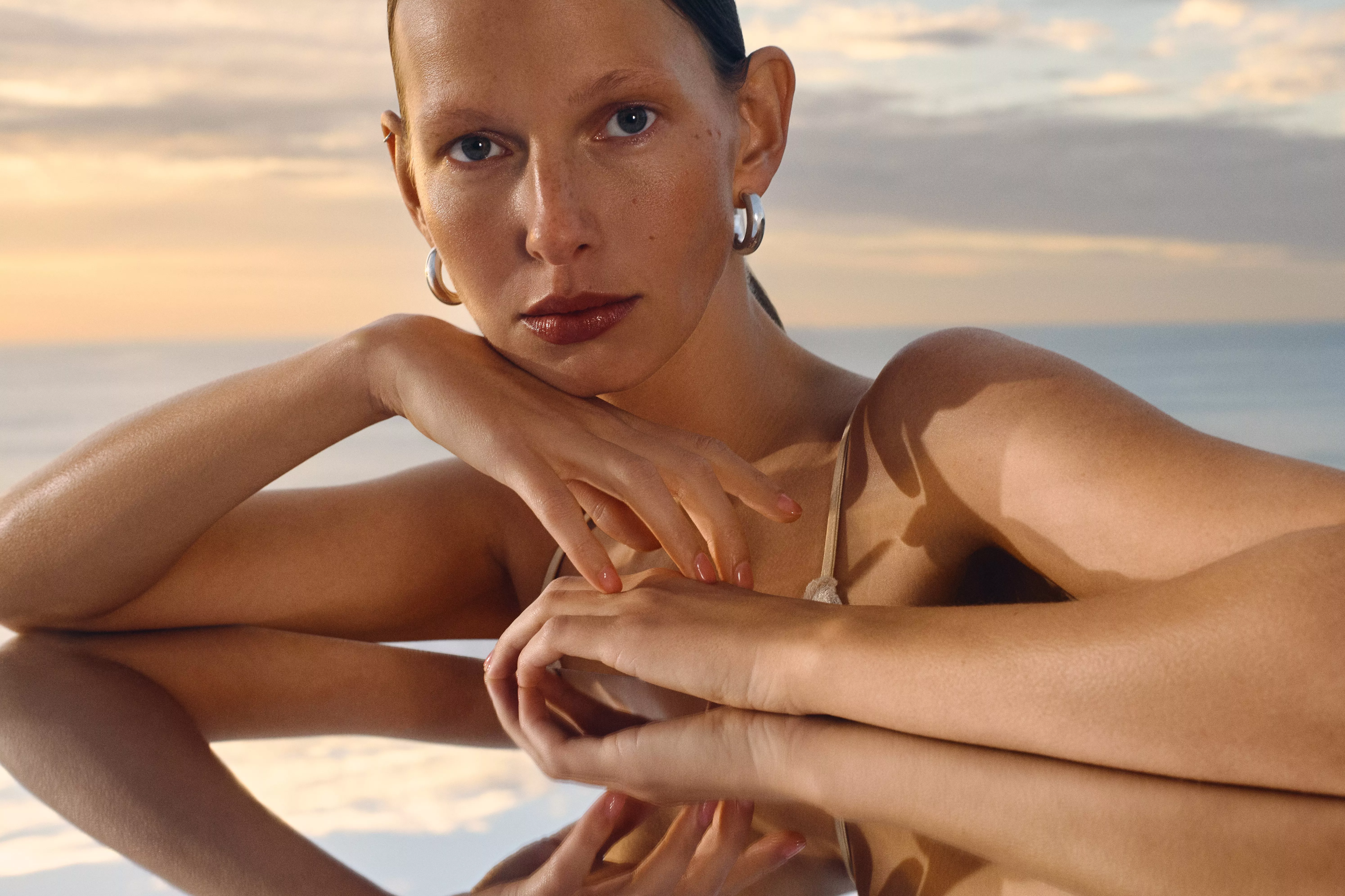 A woman with sleek hair poses against a serene sunset, wearing silver earrings and a strappy top, reflecting elegance.