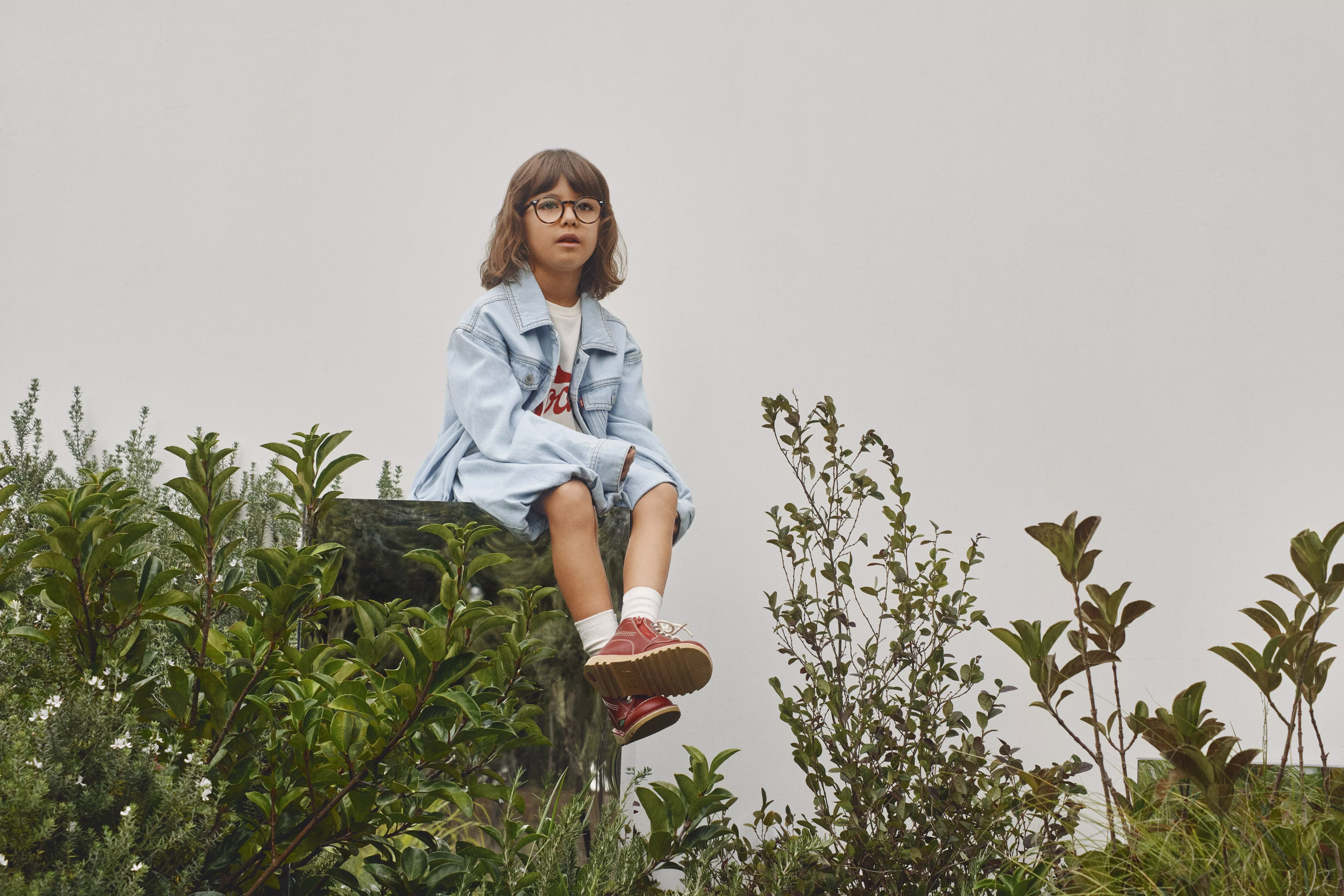 A child wearing glasses and a denim jacket sits among green plants against a plain background.