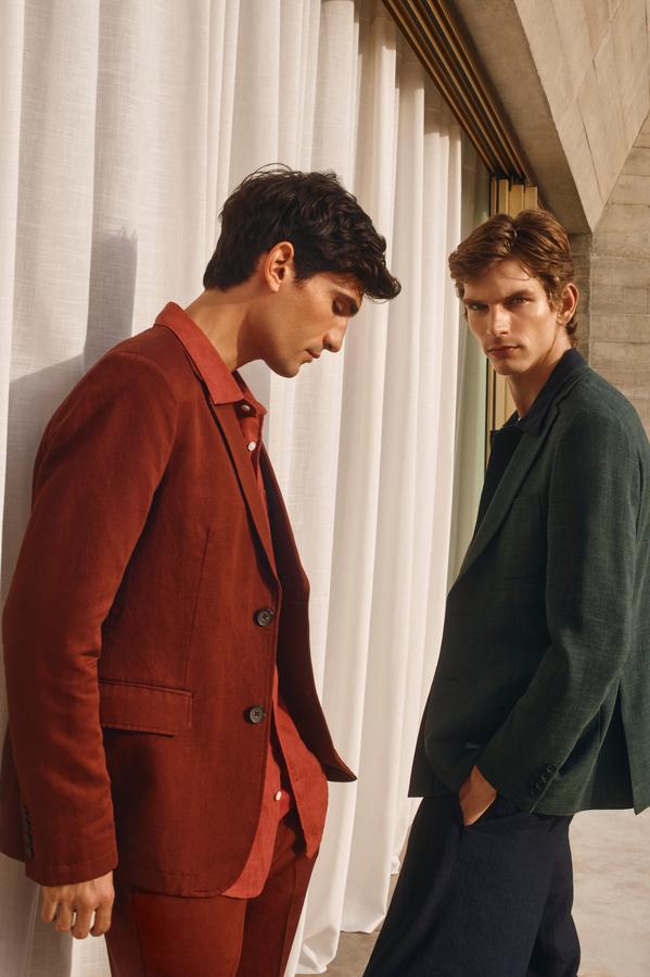 Two men in stylish suits pose indoors by white curtains, one in a rust suit, the other in dark green.
