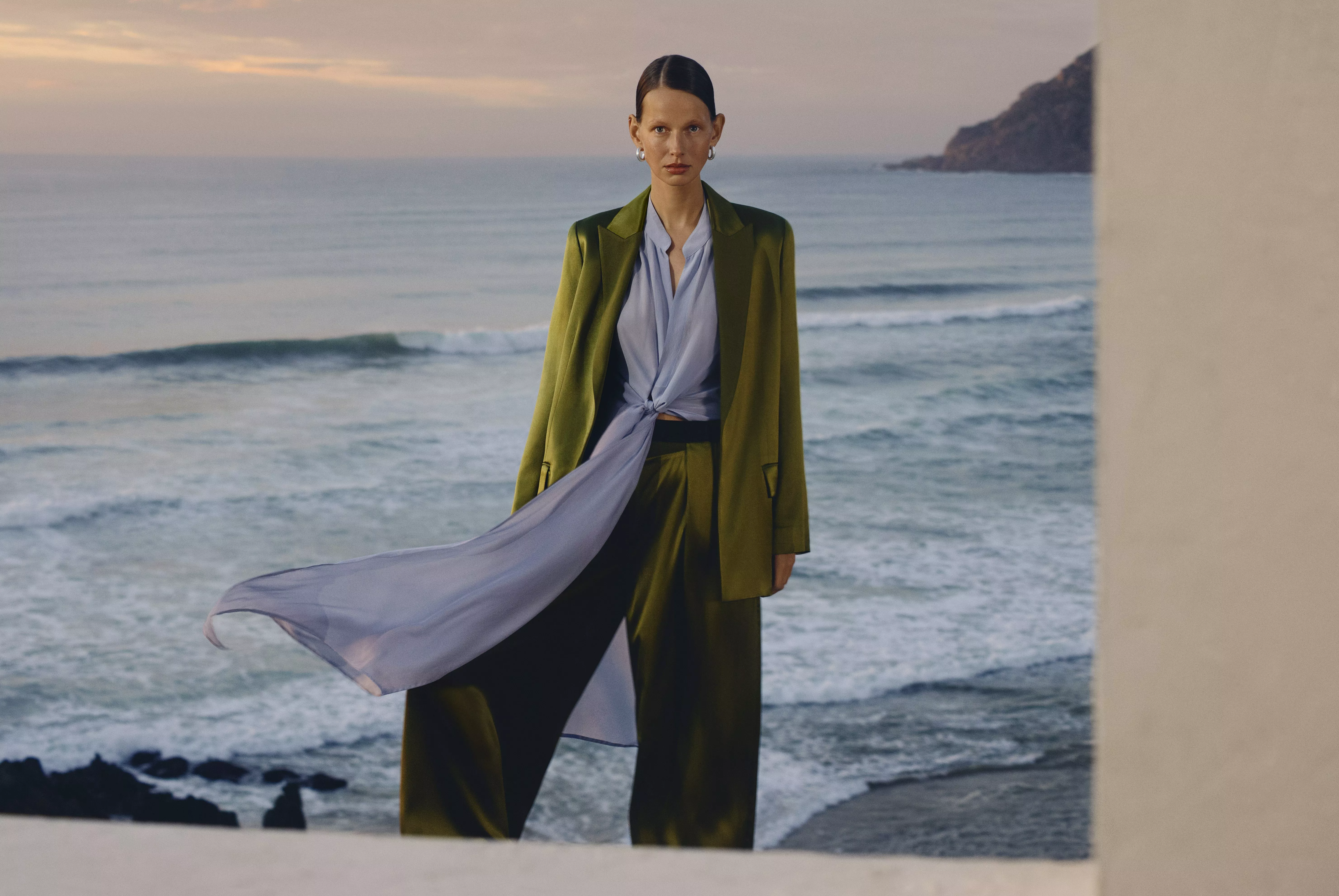 Model in a green suit and flowing shirt on coastal cliff at sunset with ocean view.