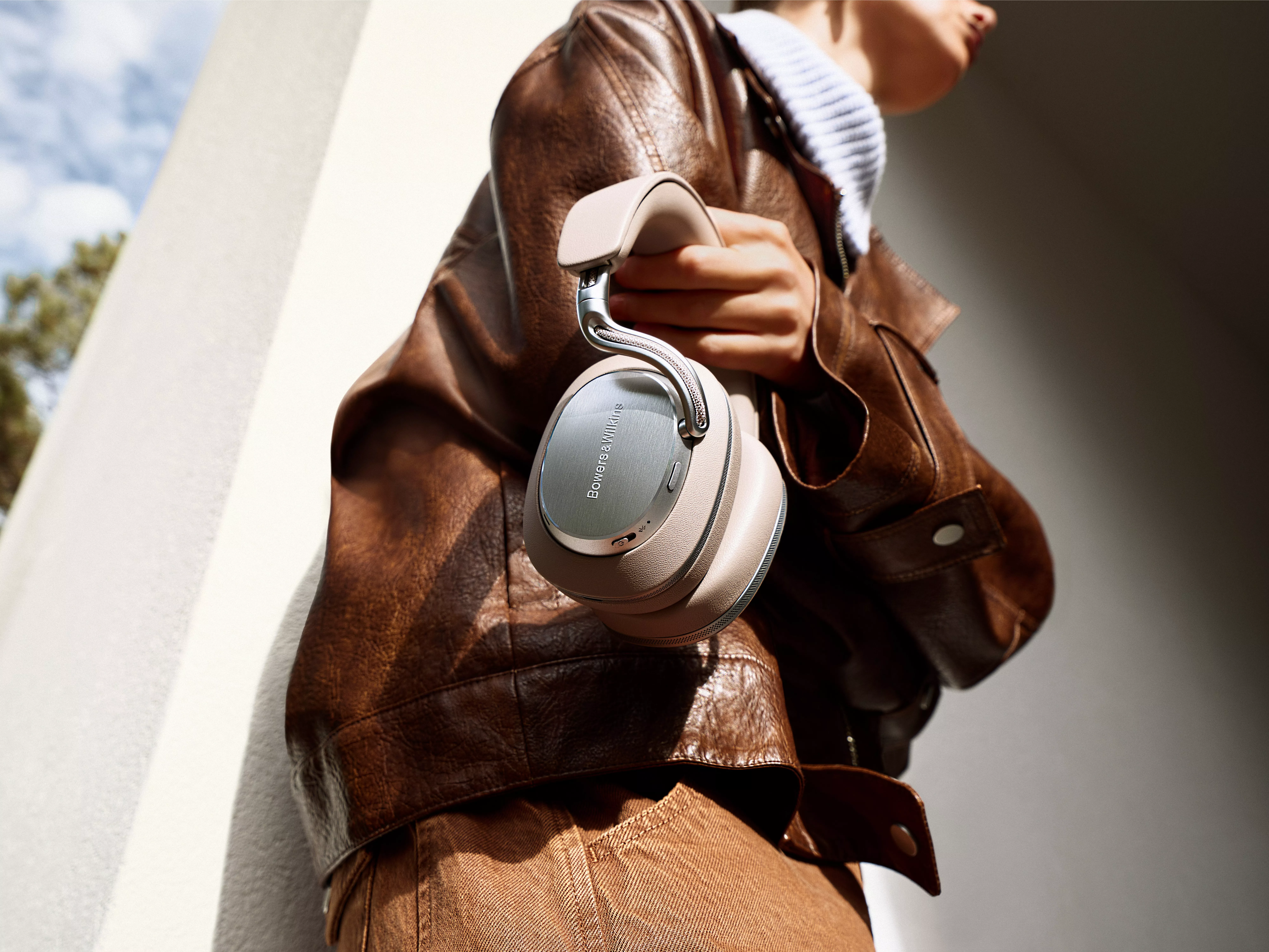 Person holds Bowers & Wilkins headphones, wearing a brown leather jacket. Bright outdoor setting. Stylish tech.
