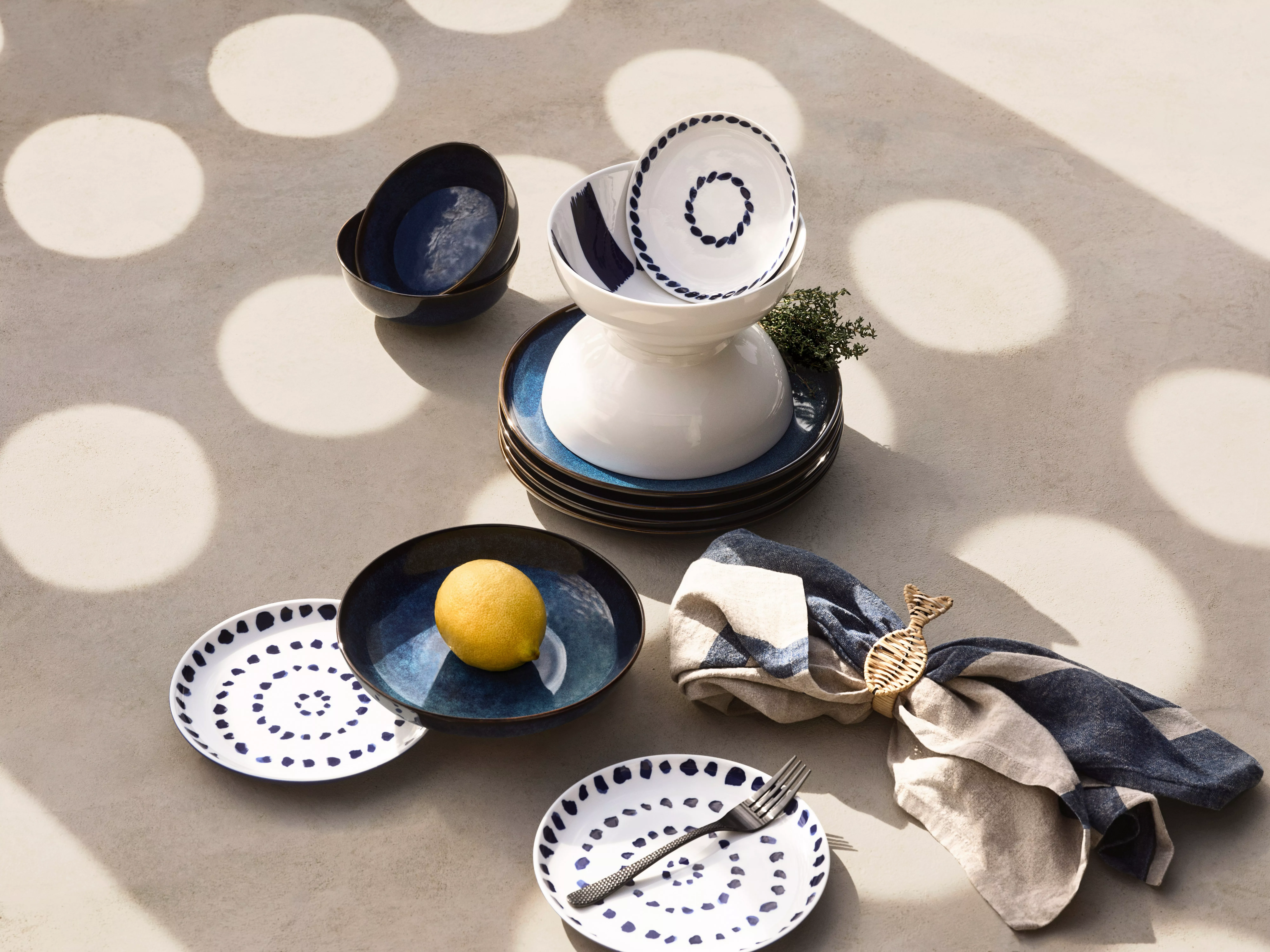 Stylish table setting with patterned plates, bowls, a lemon, and napkin under dappled sunlight on a textured surface.
