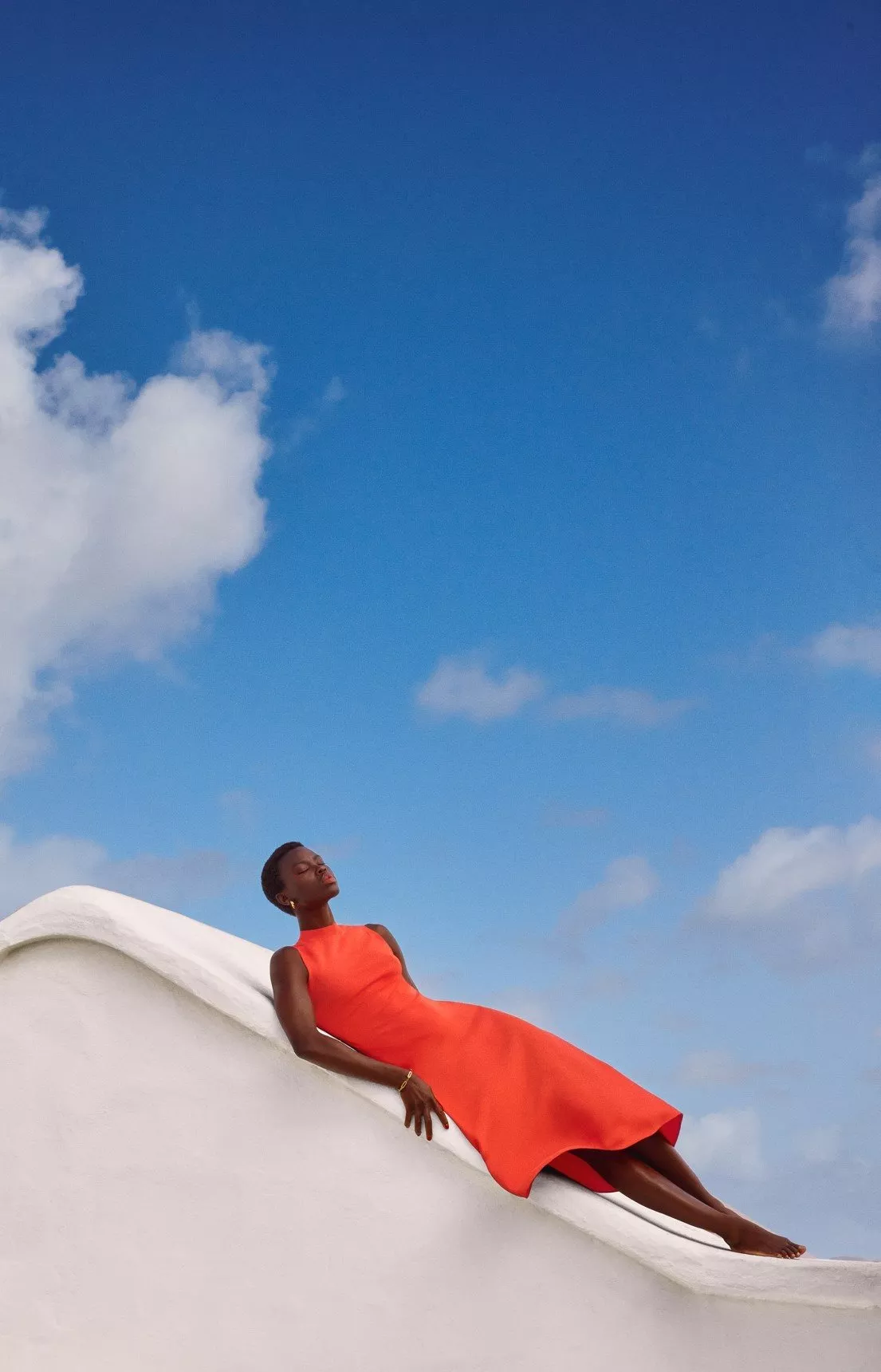 A women in a red dress laying on a white wall in front of a blue sky