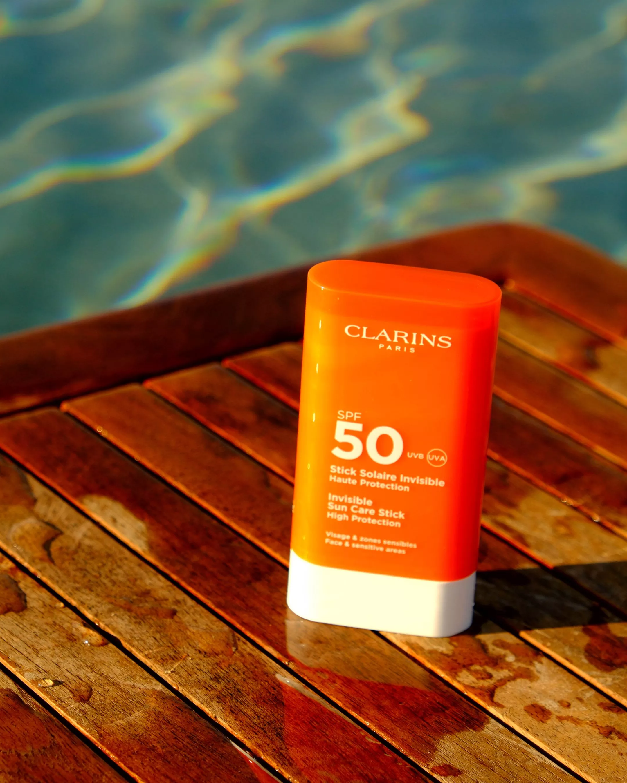Clarins SPF 50 sunscreen stick on wet wooden poolside deck with shimmering blue water reflections in background