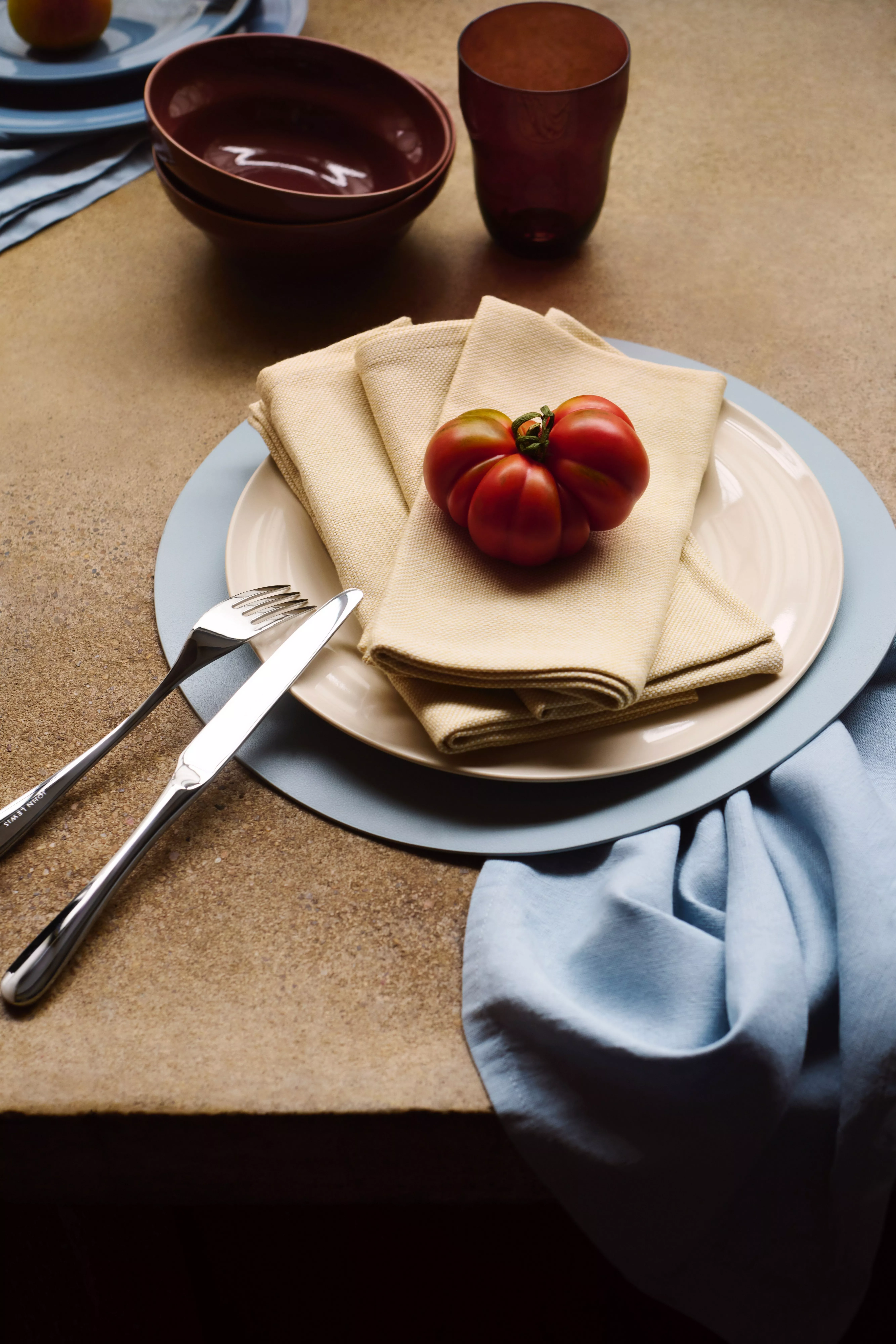 Rustic table setting with plates, linen napkin and heirloom tomato, plus cutlery, bowl and glass on stone surface
